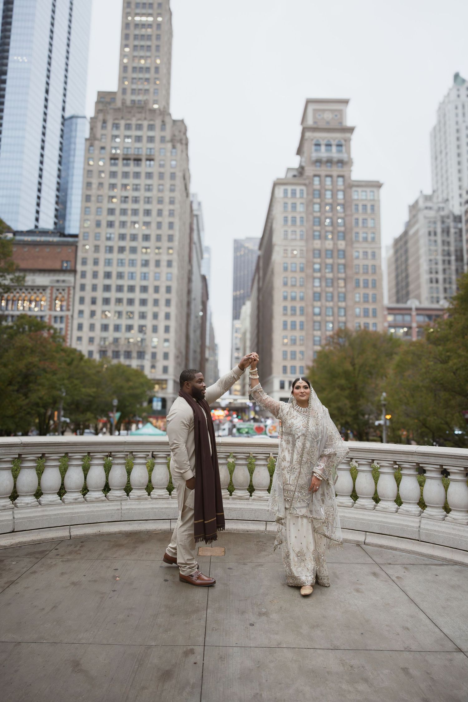 Groom spins bride in white embroidered wedding outfit on city terrace with tall buildings behind.