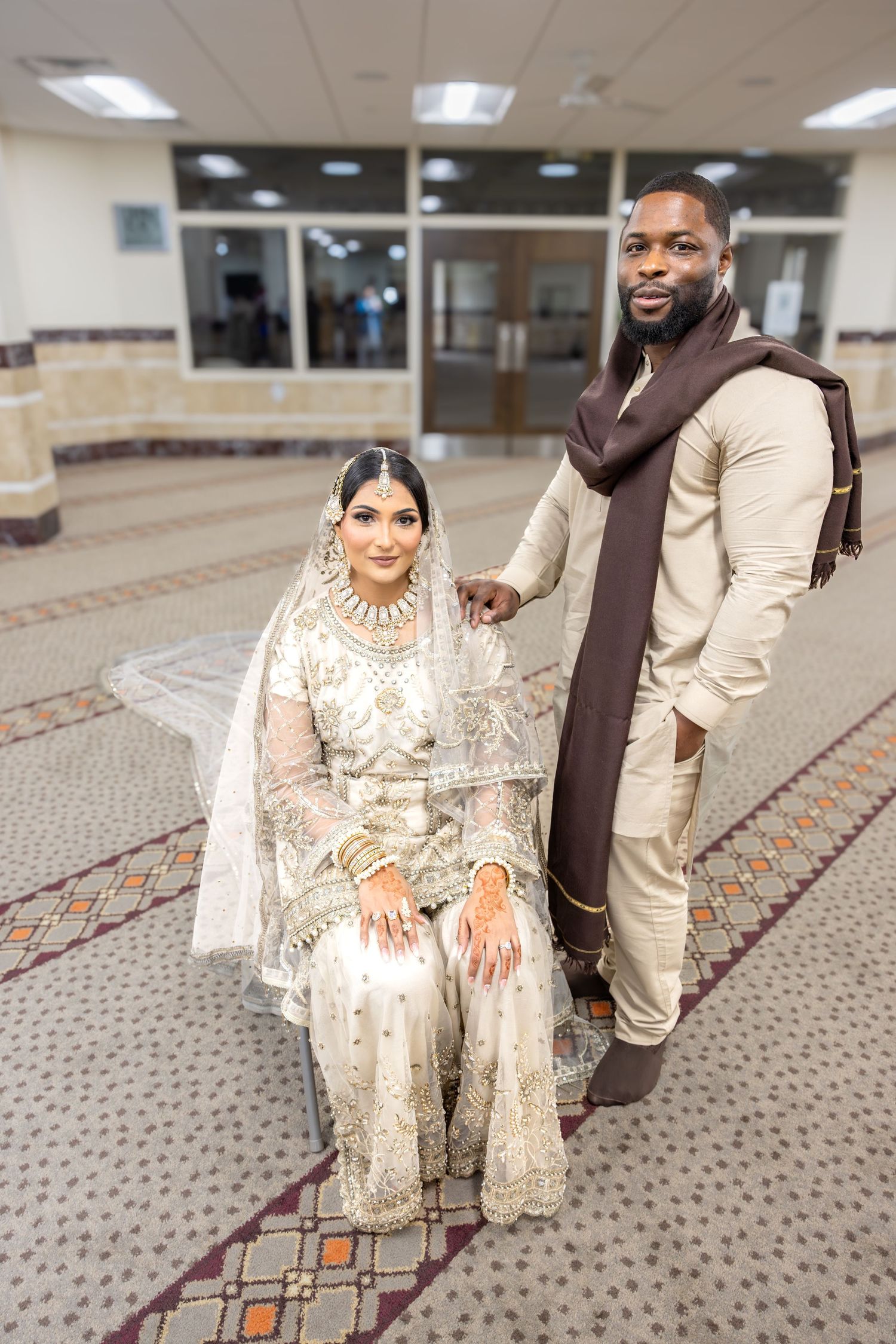 Muslim bride in white embroidered lehenga seated beside standing groom in beige sherwani indoors.