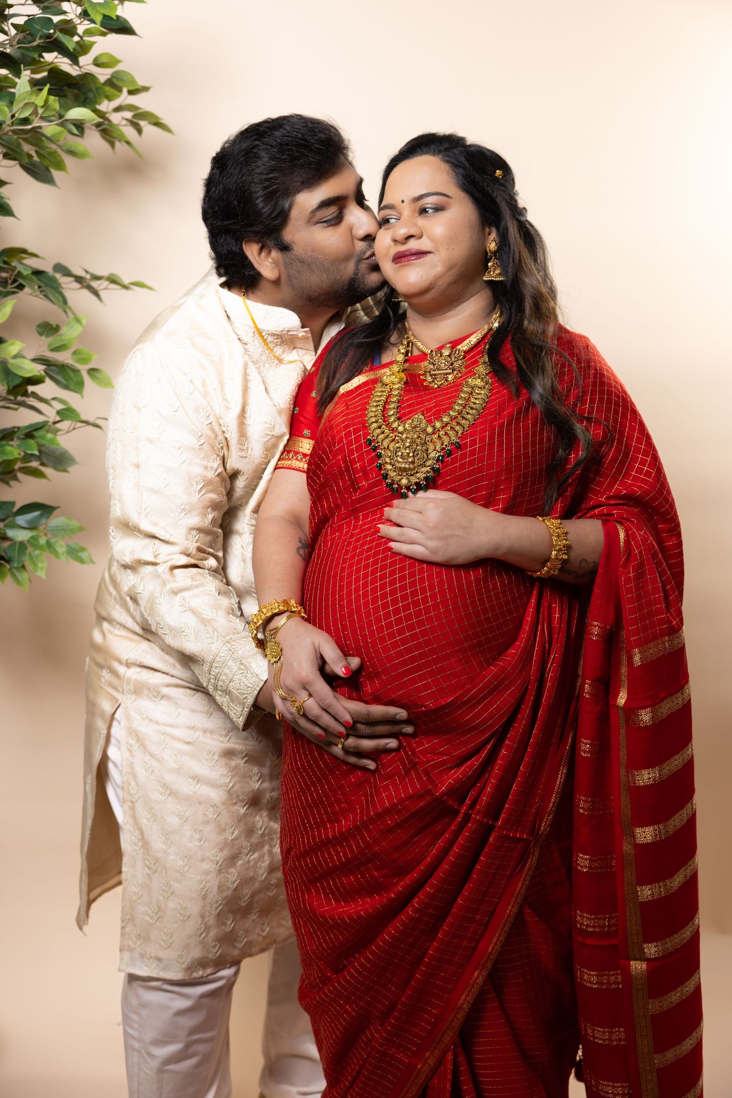Indian couple in traditional attire, pregnant woman in red saree with gold jewelry, man kissing her cheek.