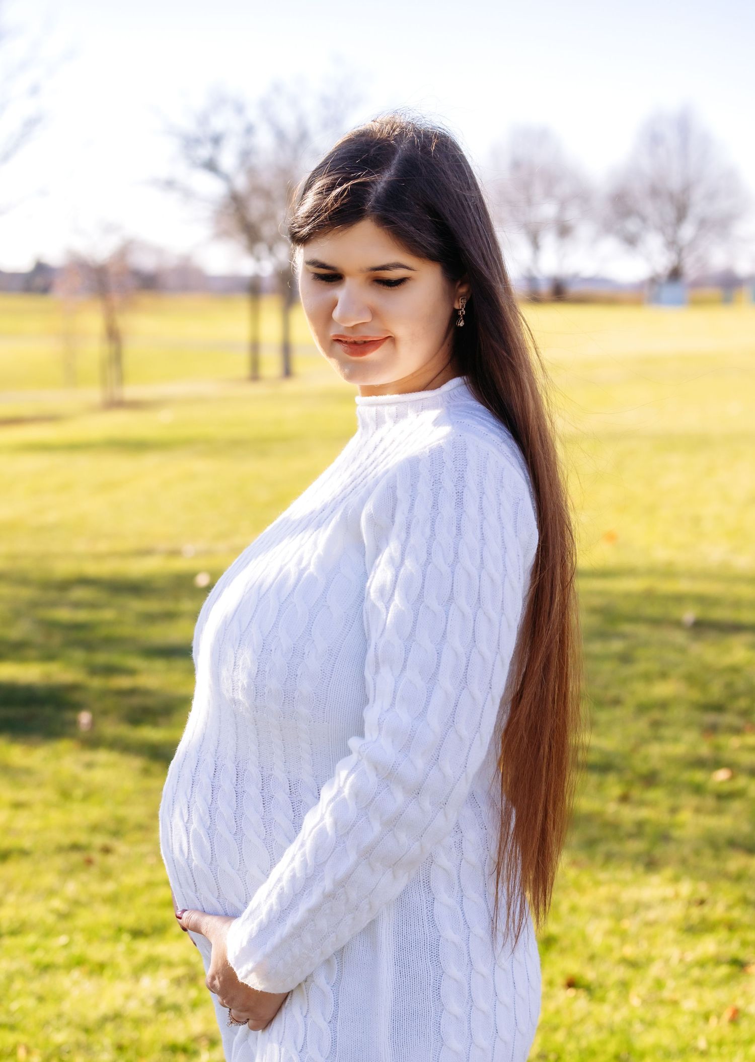 Pregnant woman in white cable-knit sweater dress cradling baby bump outdoors in sunny park.