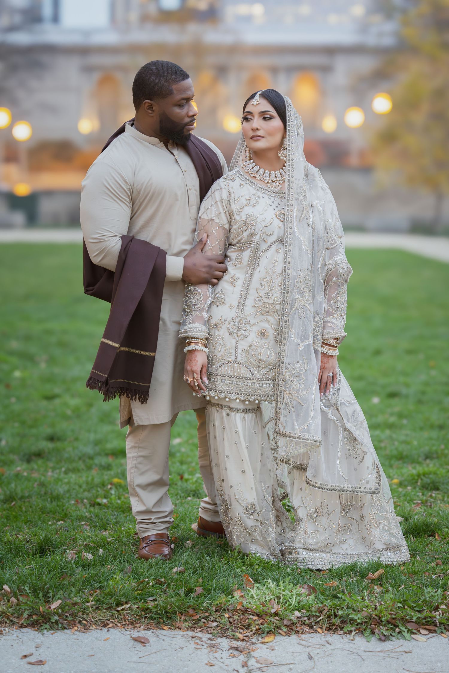 Interracial Muslim newlyweds posing outdoors, bride in ivory embroidered Pakistani bridal outfit with dupatta veil.