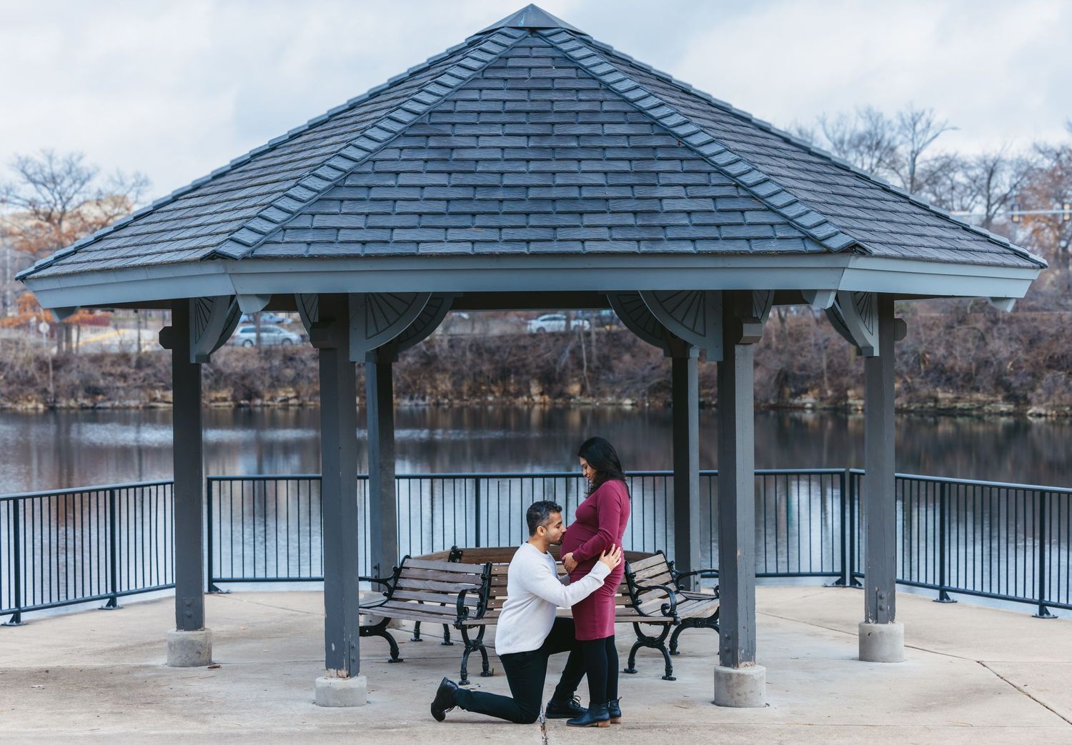 Man kneeling before pregnant woman under a gazebo by a lake in a romantic outdoor proposal.