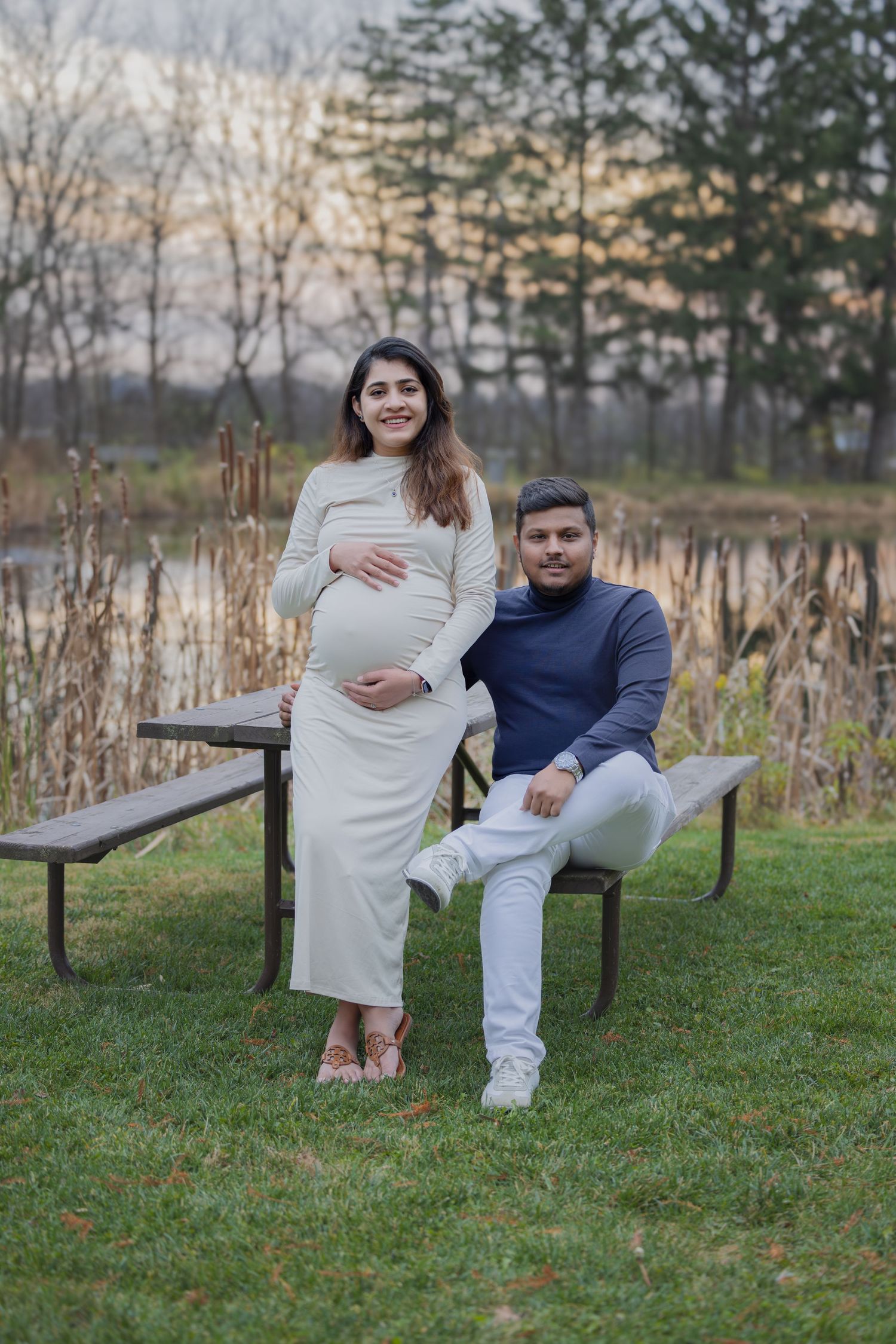 Pregnant couple posing near picnic table by a pond at dusk, woman in white dress smiling.
