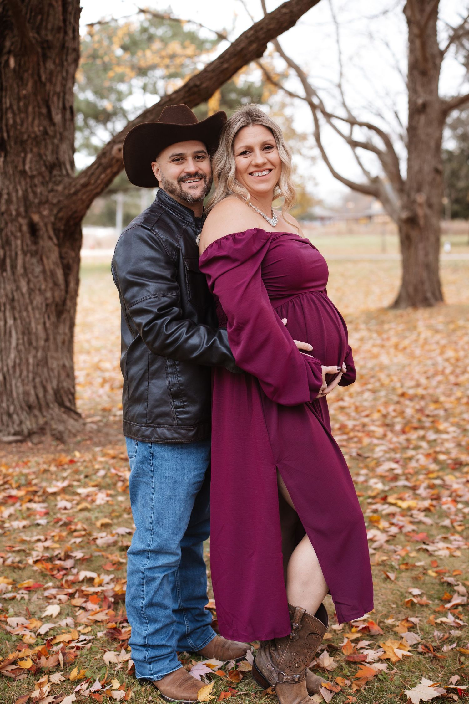 Couple posing among autumn leaves, man in cowboy hat embracing smiling pregnant woman in elegant burgundy maternity dress.