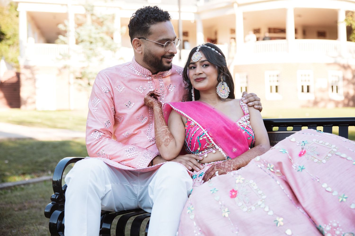 Indian wedding couple in pink traditional attire sitting on a bench smiling together.