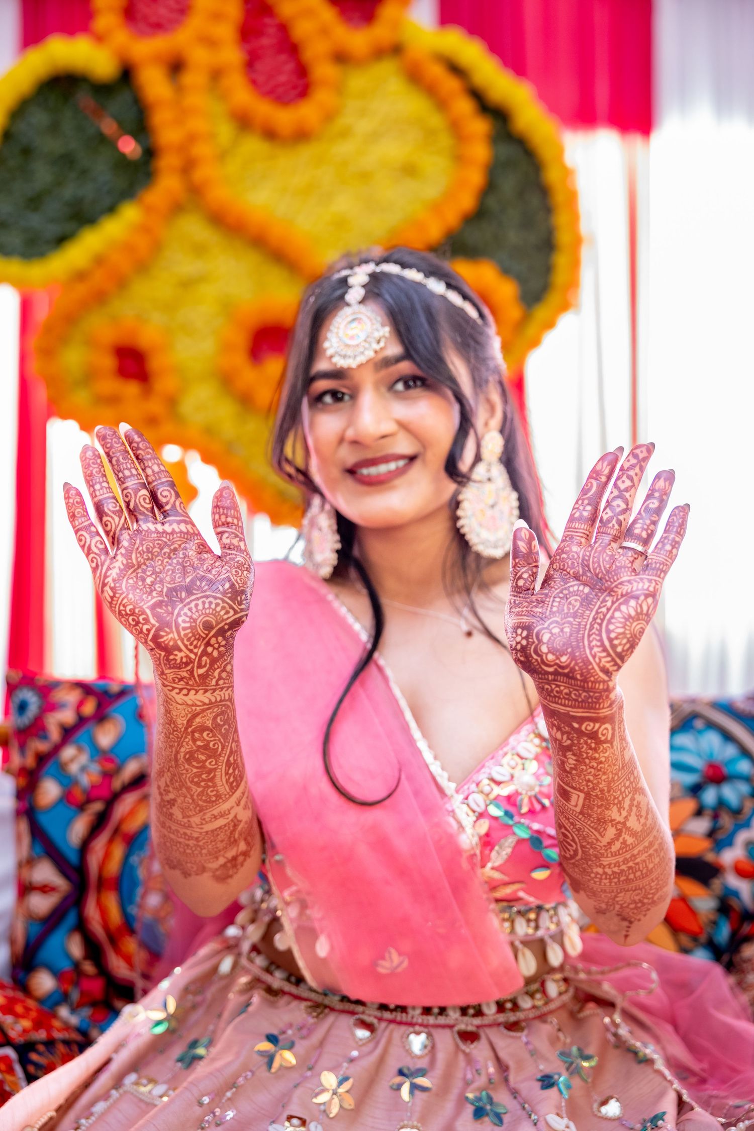 Indian bride in pink lehenga smiling and showing intricate henna mehndi designs on both hands at celebration.