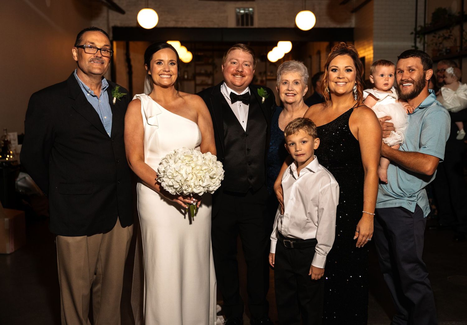 Wedding family portrait with bride, groom, and relatives at indoor reception venue.