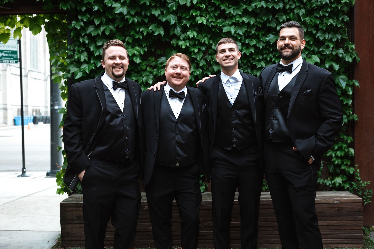 Four groomsmen in matching black tuxedos smiling together in front of ivy-covered wall.