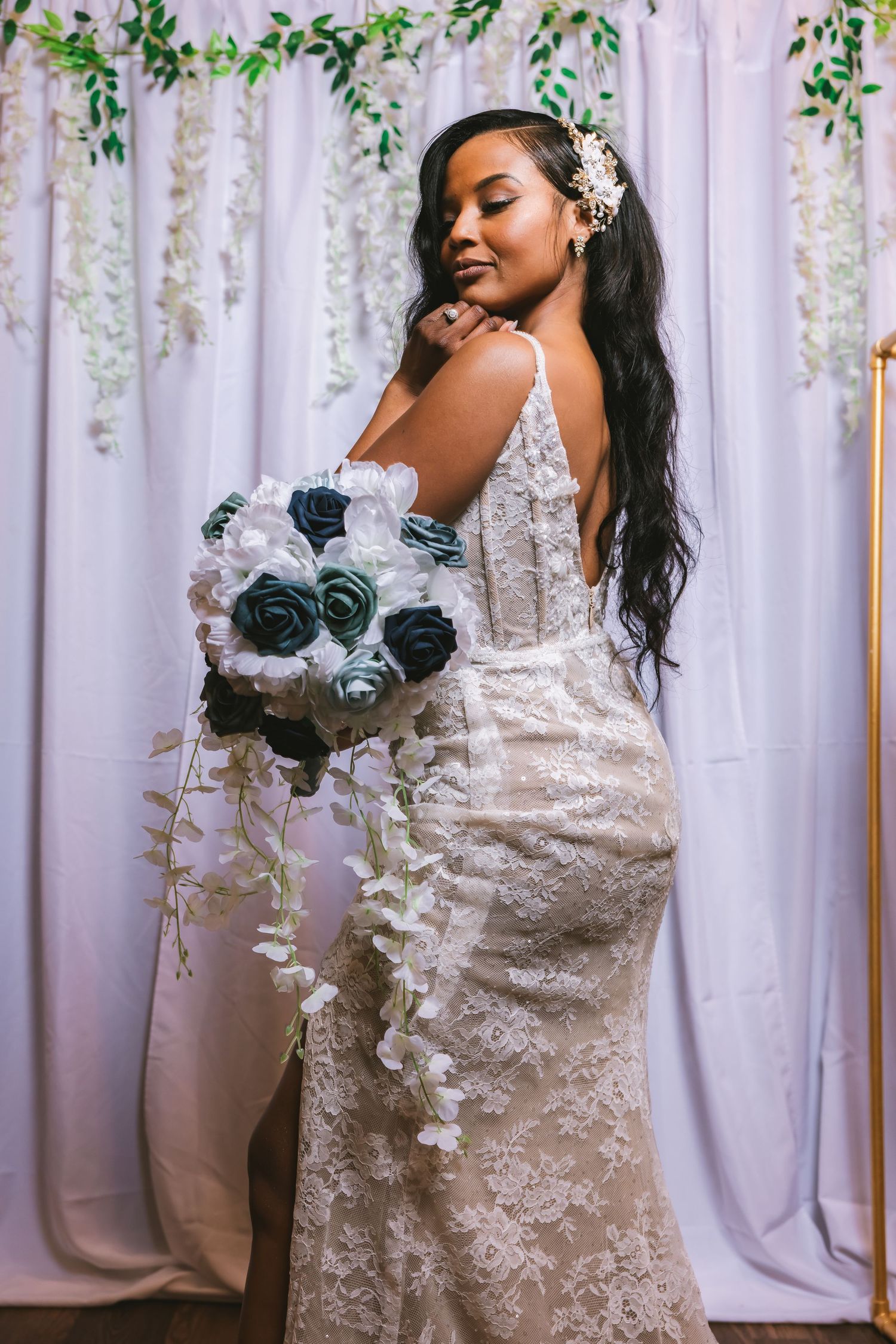 A bride in a backless lace gown poses with a blue and white cascading bouquet against white draping.