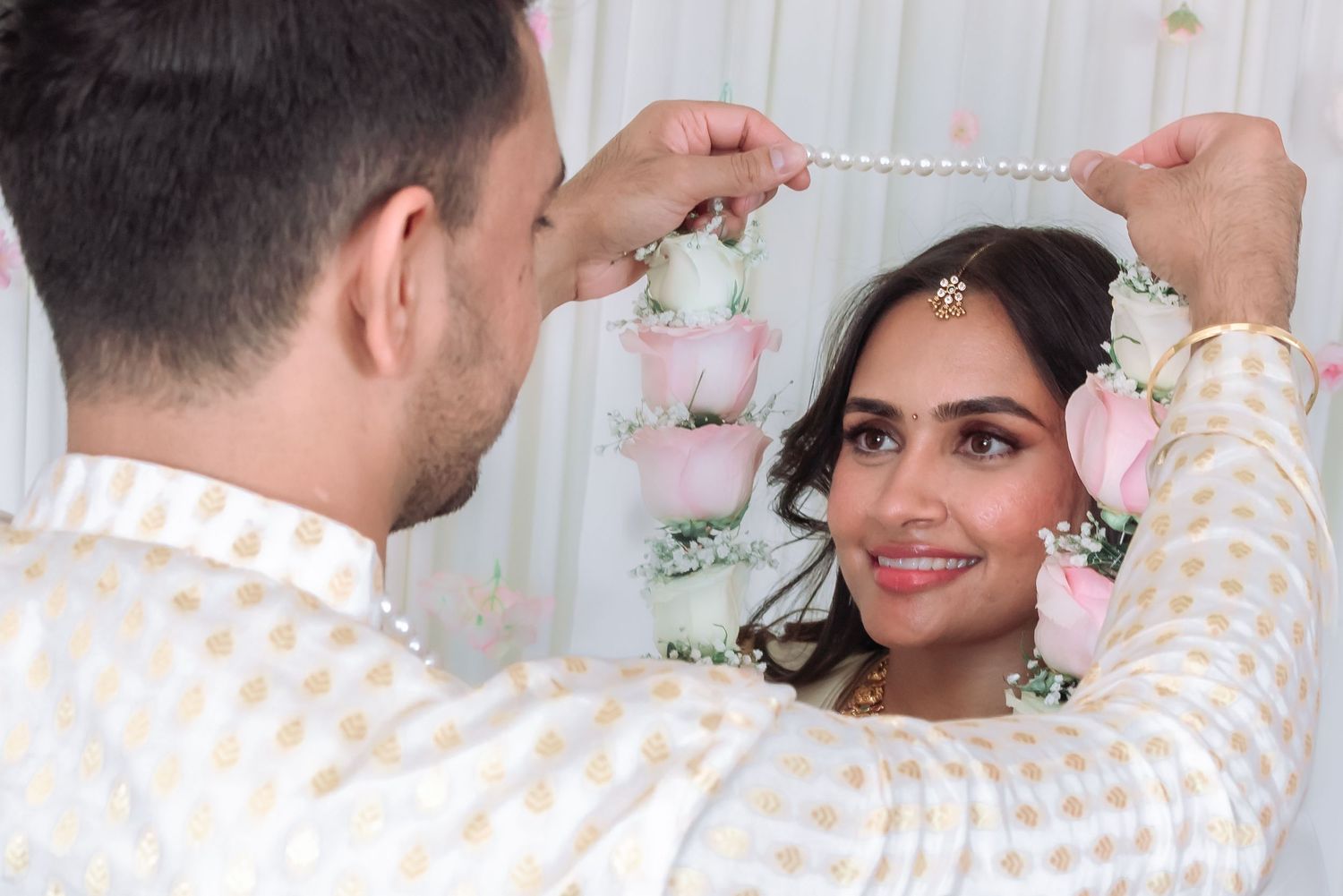Groom placing a pearl necklace on smiling bride during traditional Indian wedding ceremony with floral decorations.