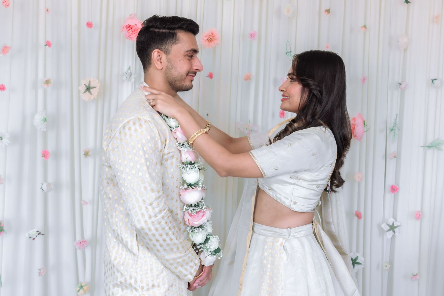 Indian couple smiling as bride places floral garland around grooms neck during wedding ceremony.