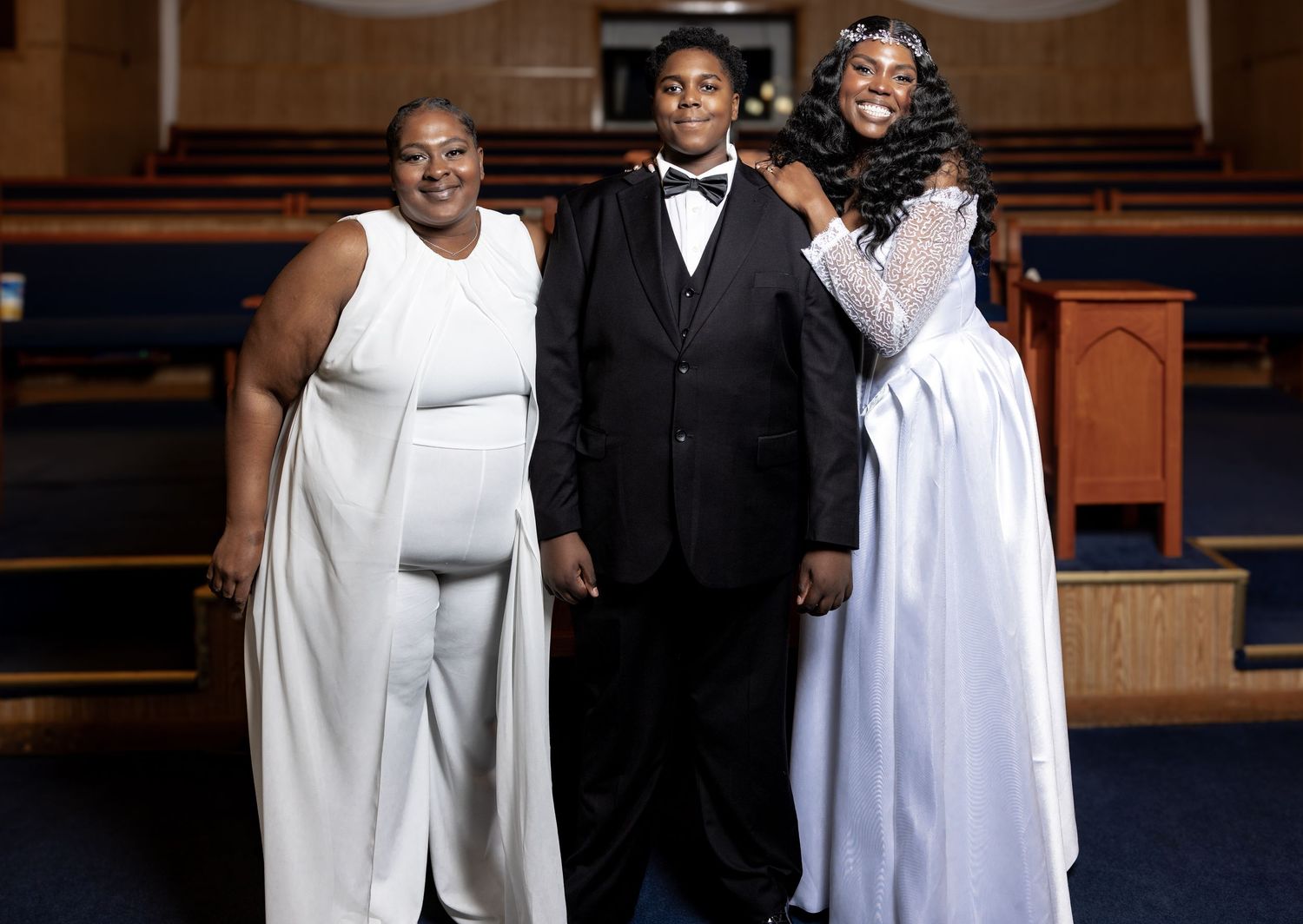 Three people in white and black formal wear posing together inside a church with wooden pews.