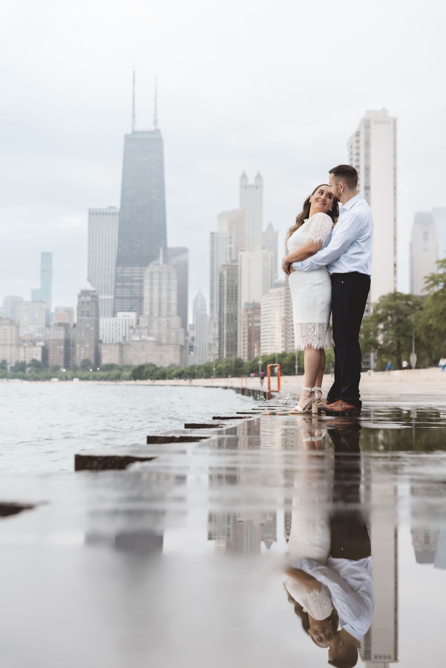 Couple embracing on Chicago lakefront with city skyline reflection in water on a cloudy day.