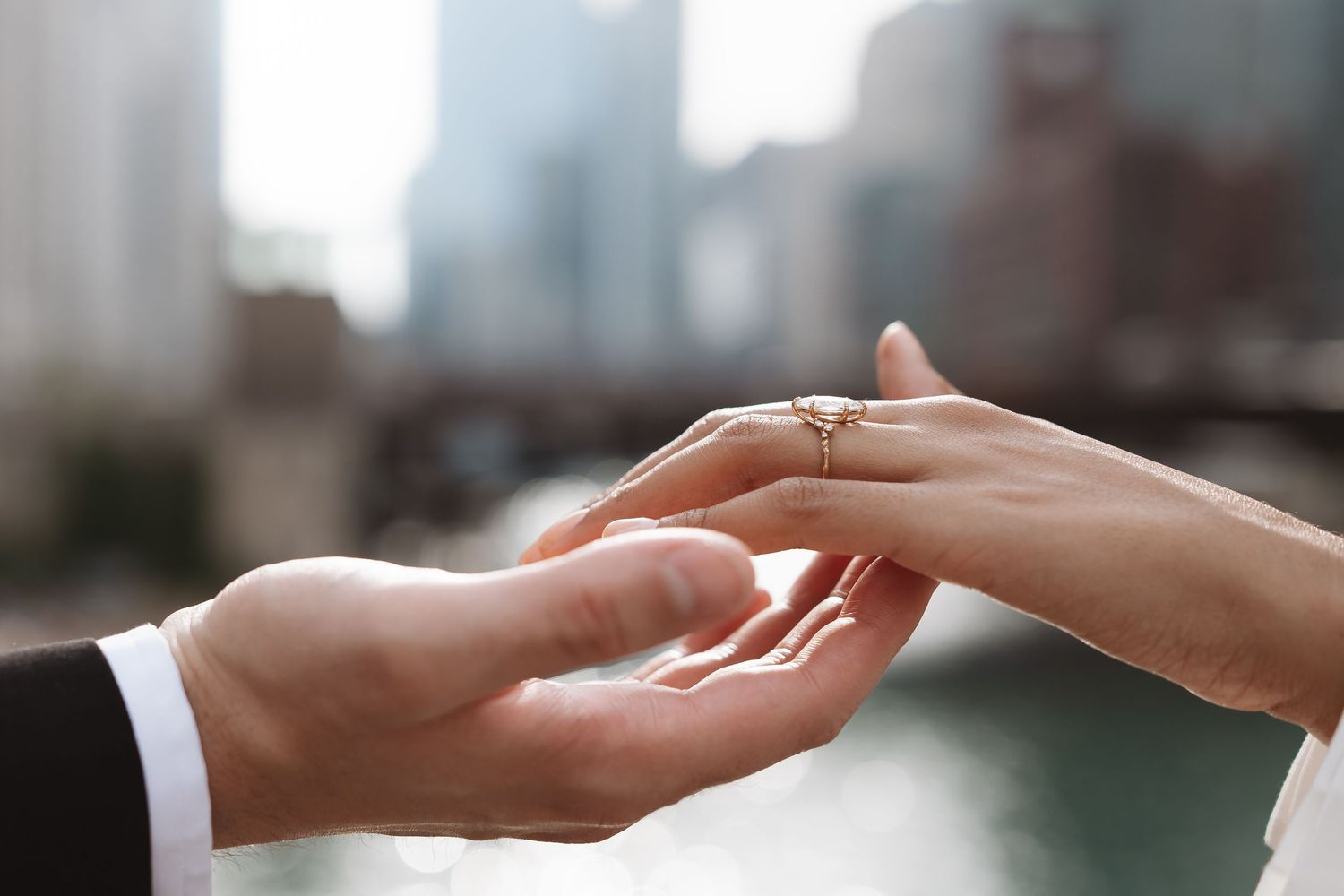 Close-up of a man placing a diamond engagement ring on a woman's finger by a city river.