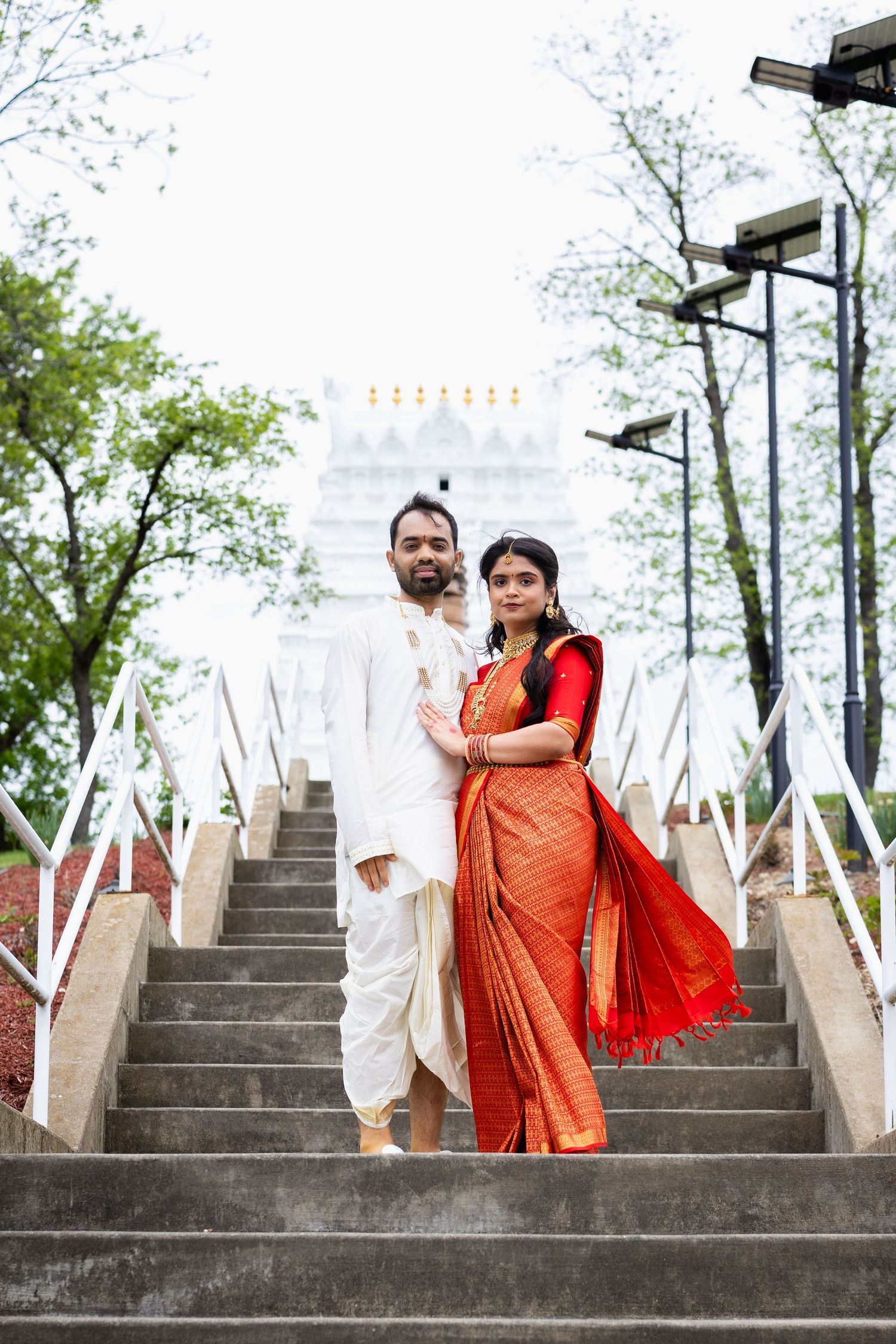 Indian wedding couple in traditional attire posing on temple steps with white architecture behind them.