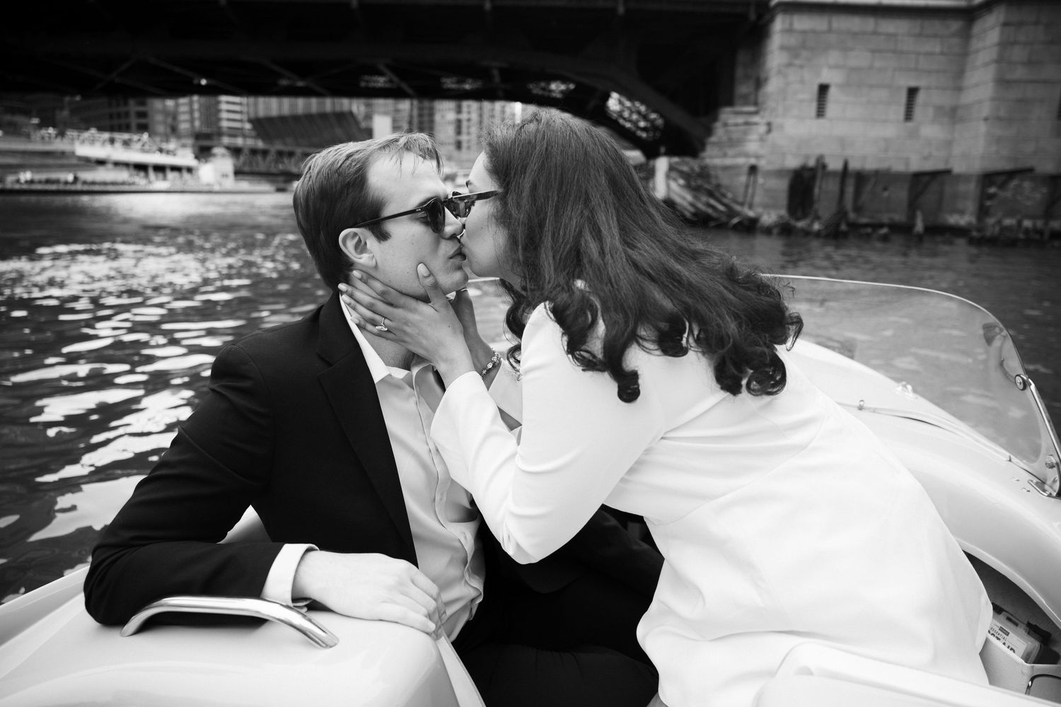 Black and white photo of a couple sharing a romantic kiss on a boat under a city bridge.