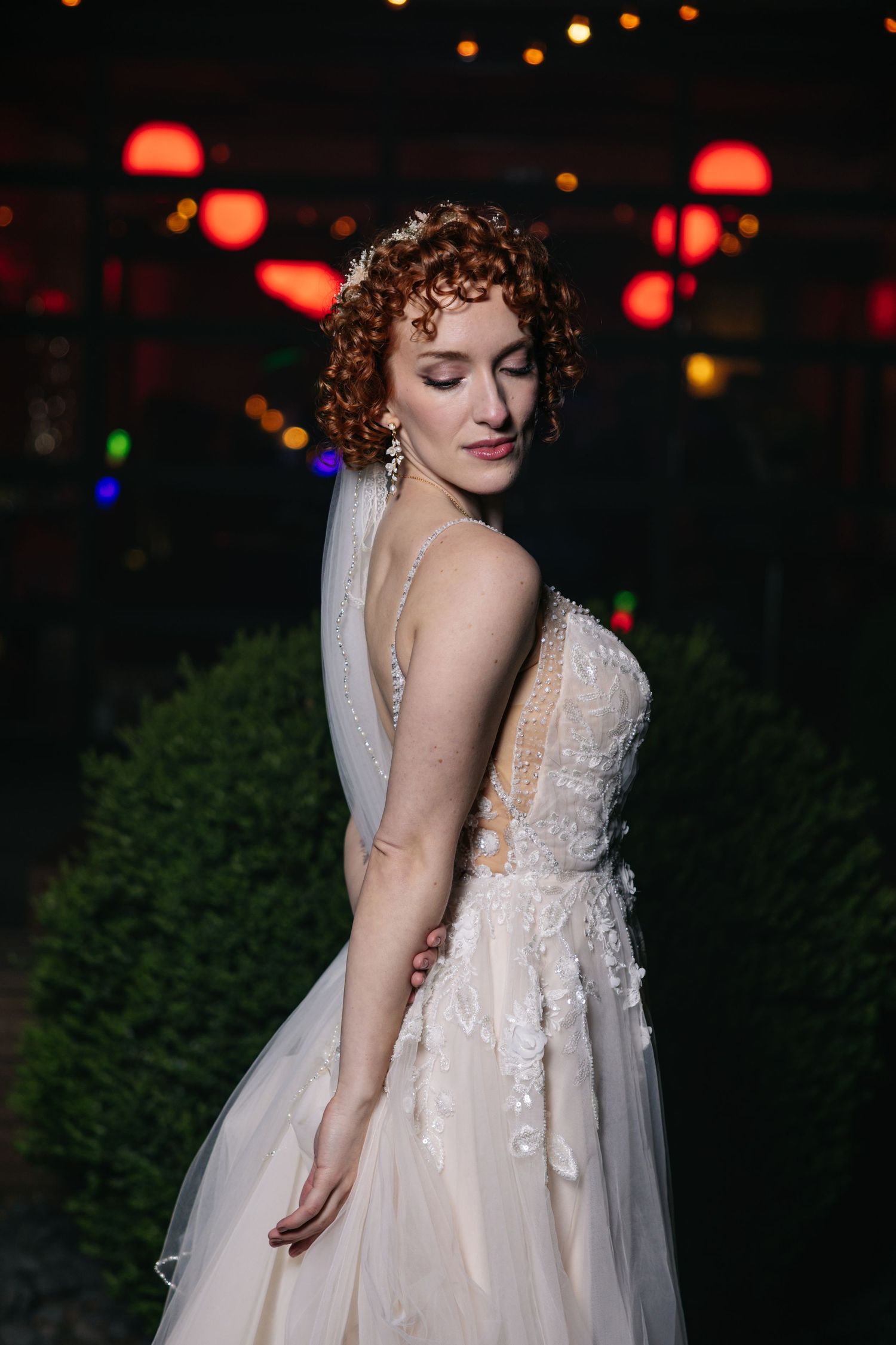 Bride in lace wedding gown with veil poses outdoors at night with red lanterns glowing in background.