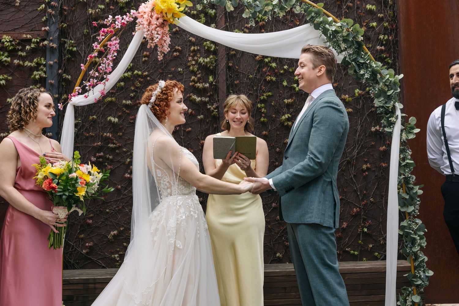 Bride and groom exchange vows under floral arch with bridesmaid and officiant at outdoor wedding ceremony.