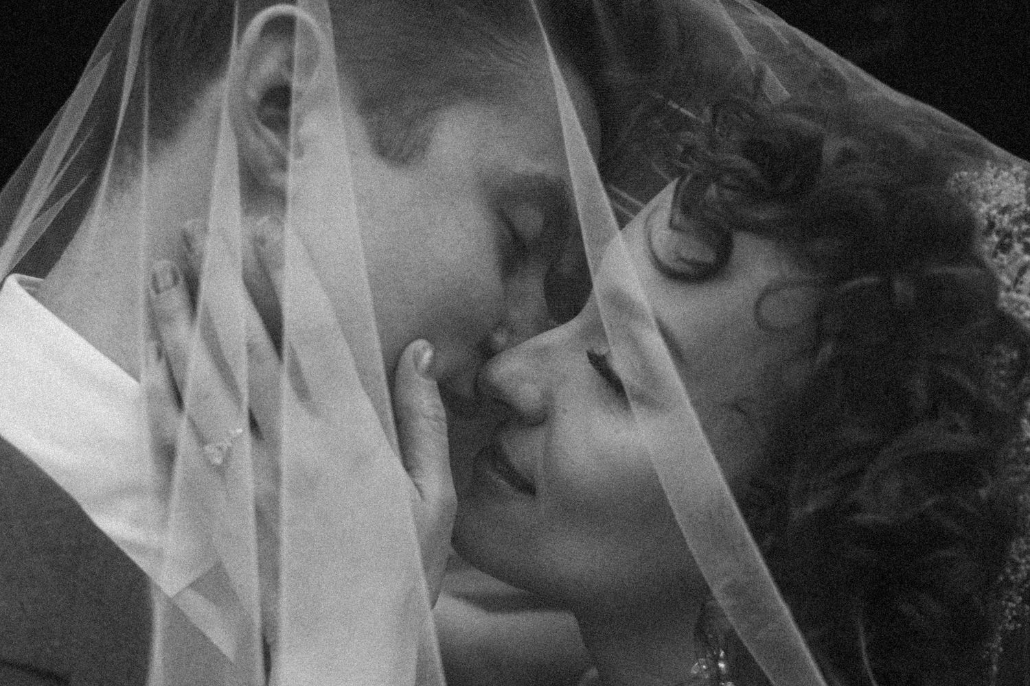 Black and white photo of bride and groom sharing a romantic kiss under a wedding veil.