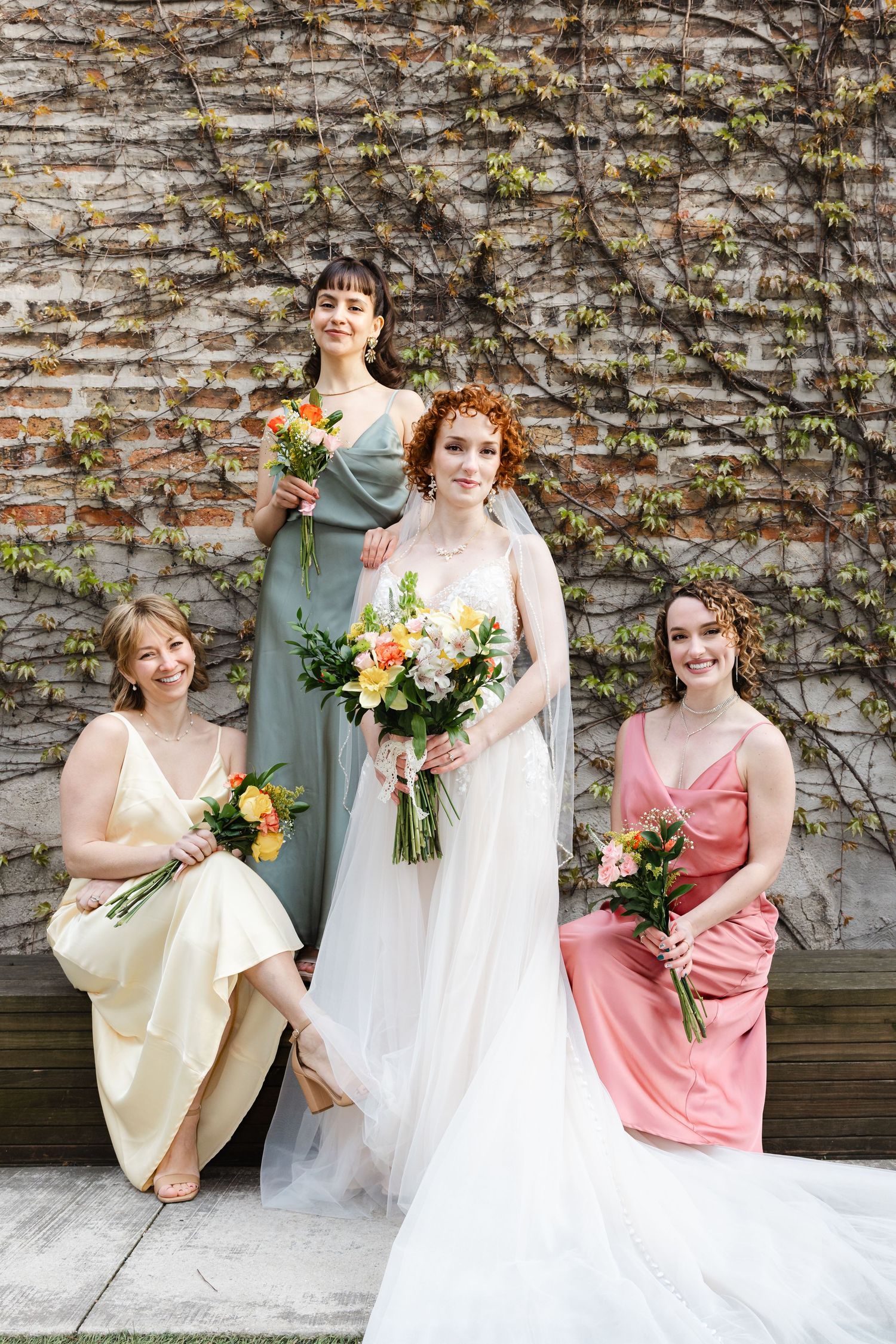 Bride in white gown poses with three bridesmaids in colorful dresses holding bouquets against a brick wall.