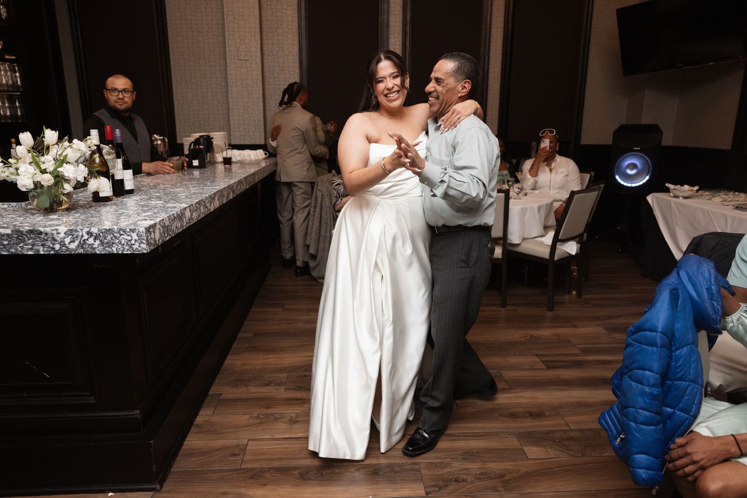 Bride in white satin gown shares a dance with a man in gray suit at wedding reception.