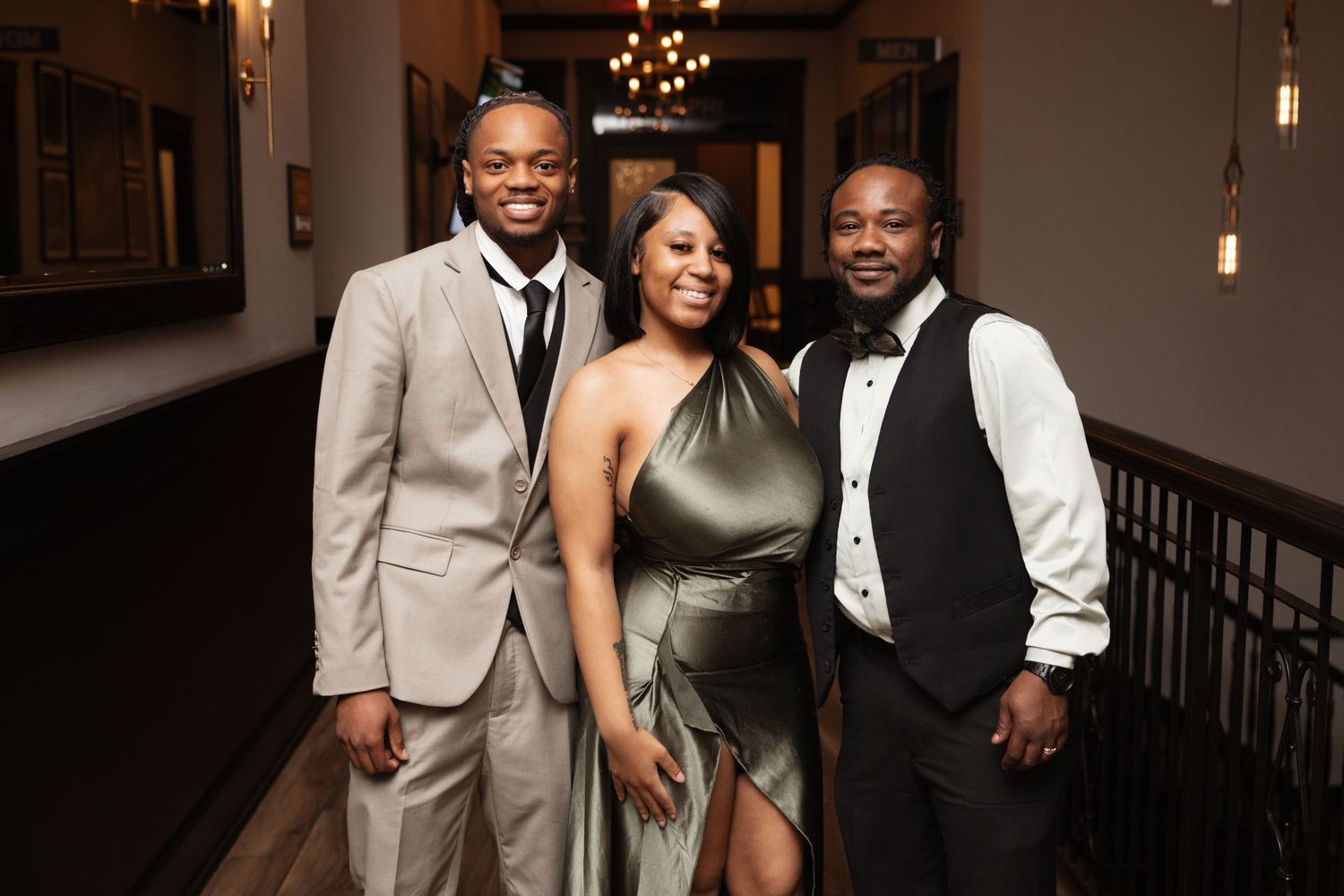 Three formally dressed friends smiling together in elegant venue, wearing gray suit, green gown, and tuxedo vest.