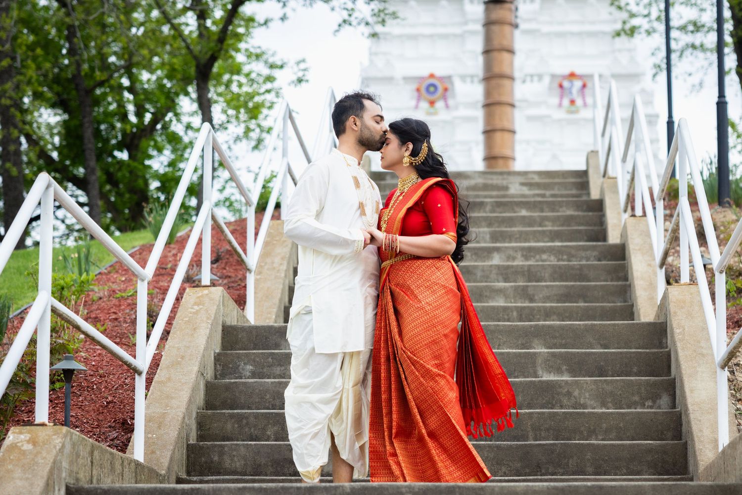 Indian couple in traditional wedding attire sharing a kiss on outdoor stone steps.