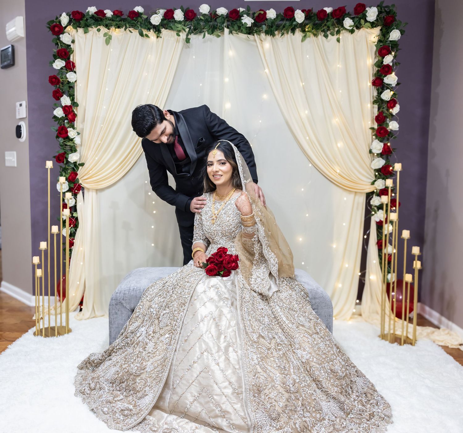 Groom leans toward seated bride in ornate gold lehenga beneath floral wedding arch with string lights.