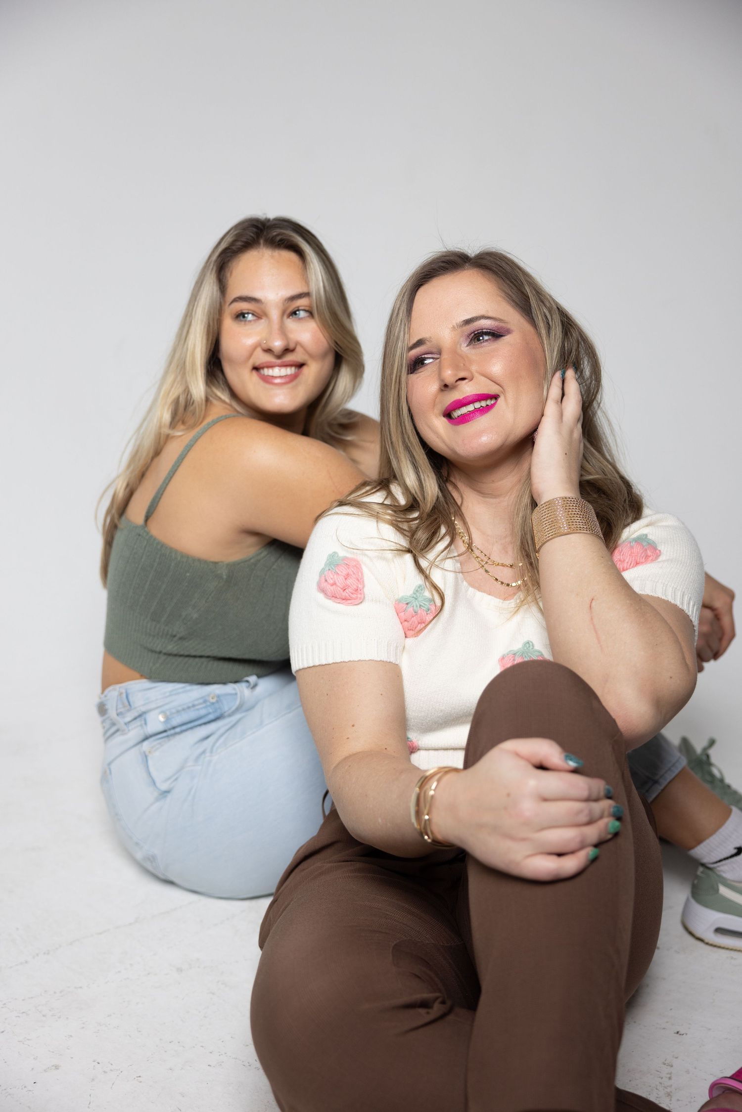 Two smiling blonde women sitting back-to-back on white floor in colorful casual outfits in studio.