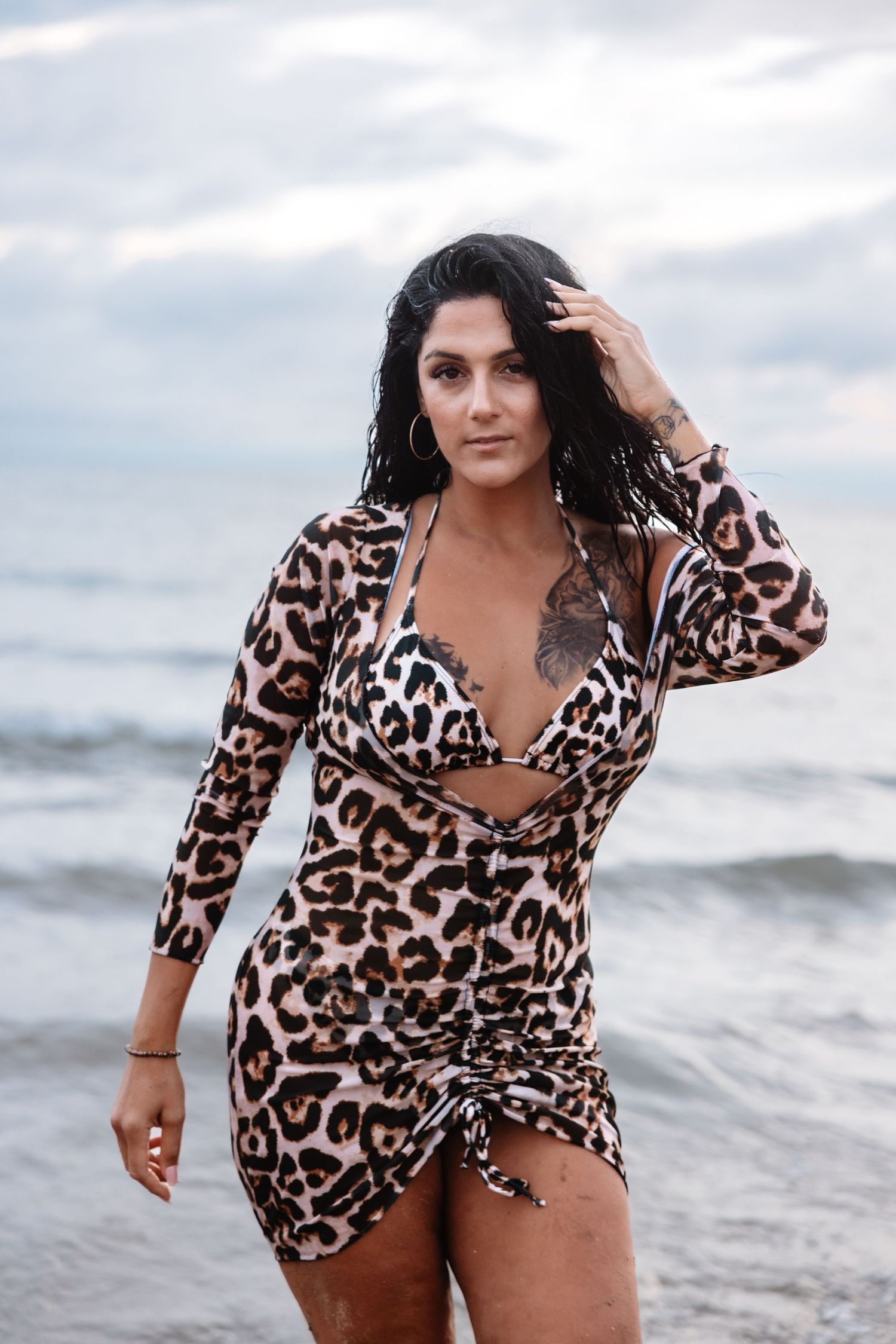 Woman with tattoo in leopard print dress touching wet hair while standing on beach at dusk.