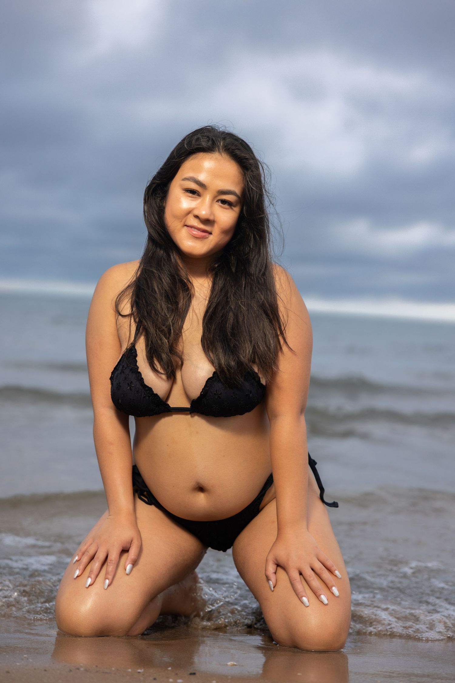 Young woman in black bikini kneeling on wet sandy beach with ocean waves and cloudy sky behind her.