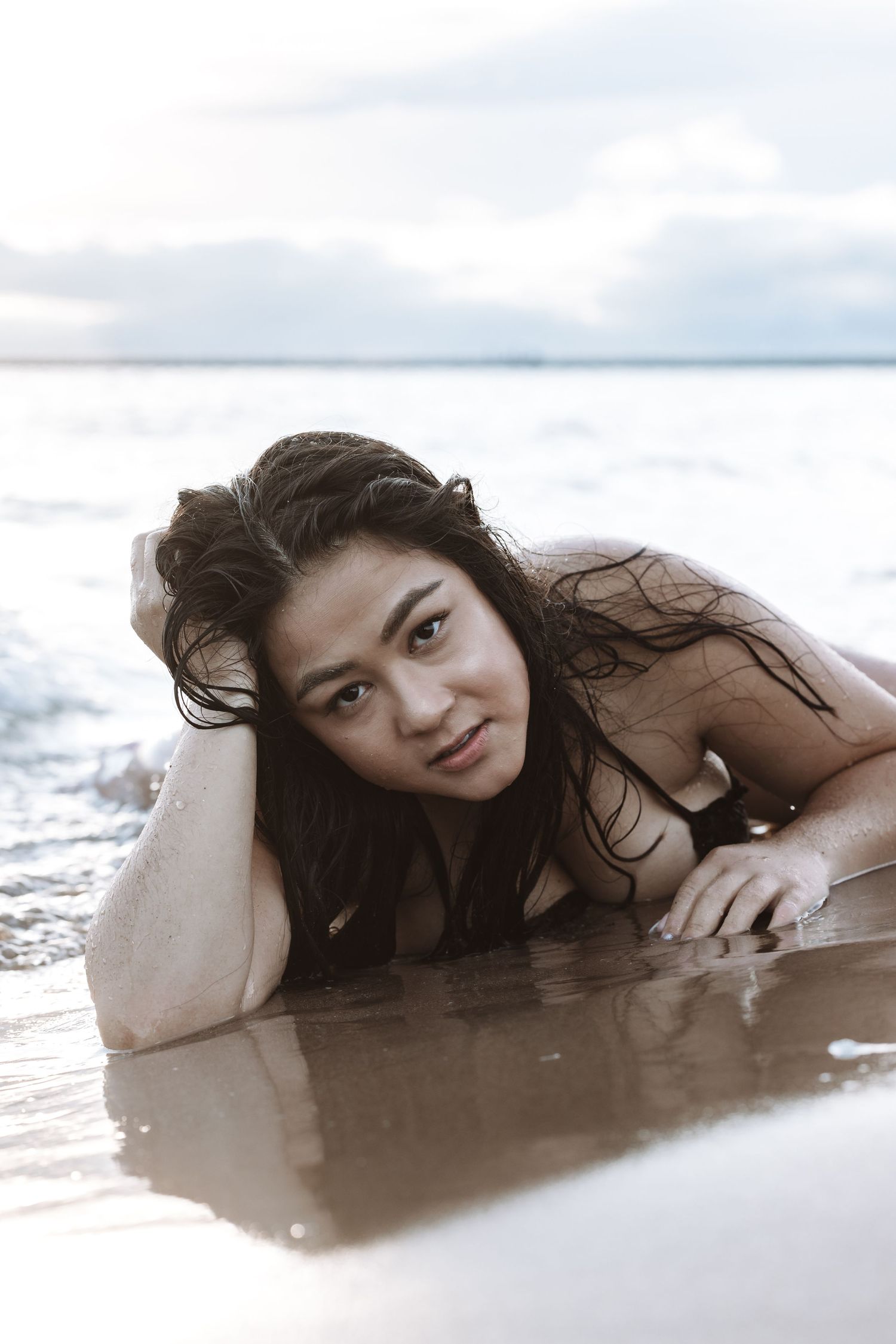 Woman with long wet hair lying on wet sand in shallow ocean waves at beach.