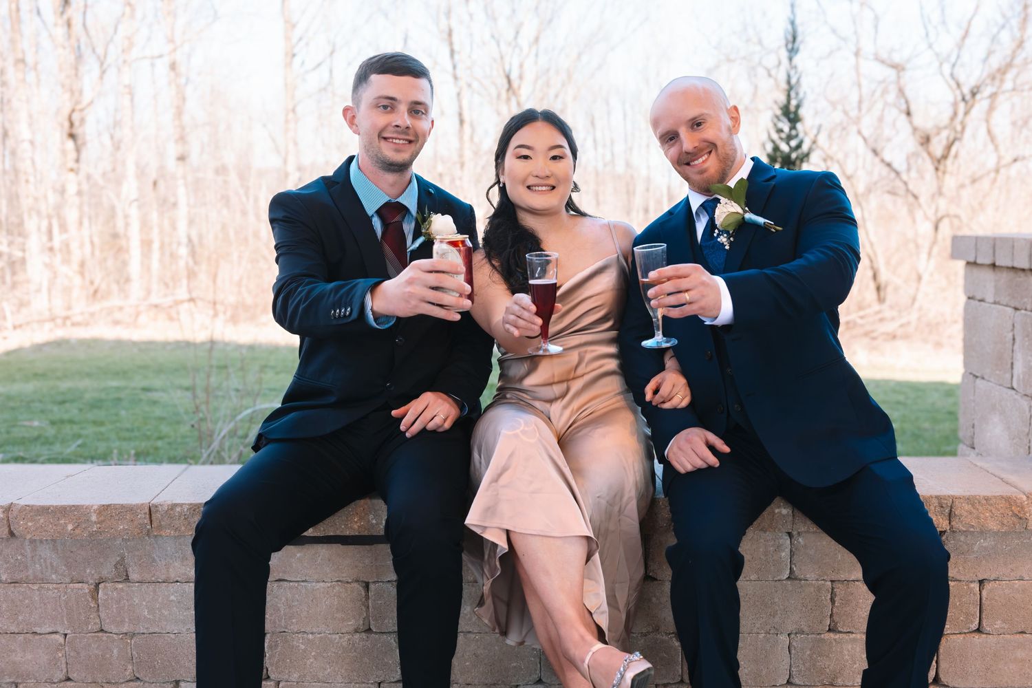 Three wedding guests in formal attire toasting with champagne glasses outdoors at a wedding reception.