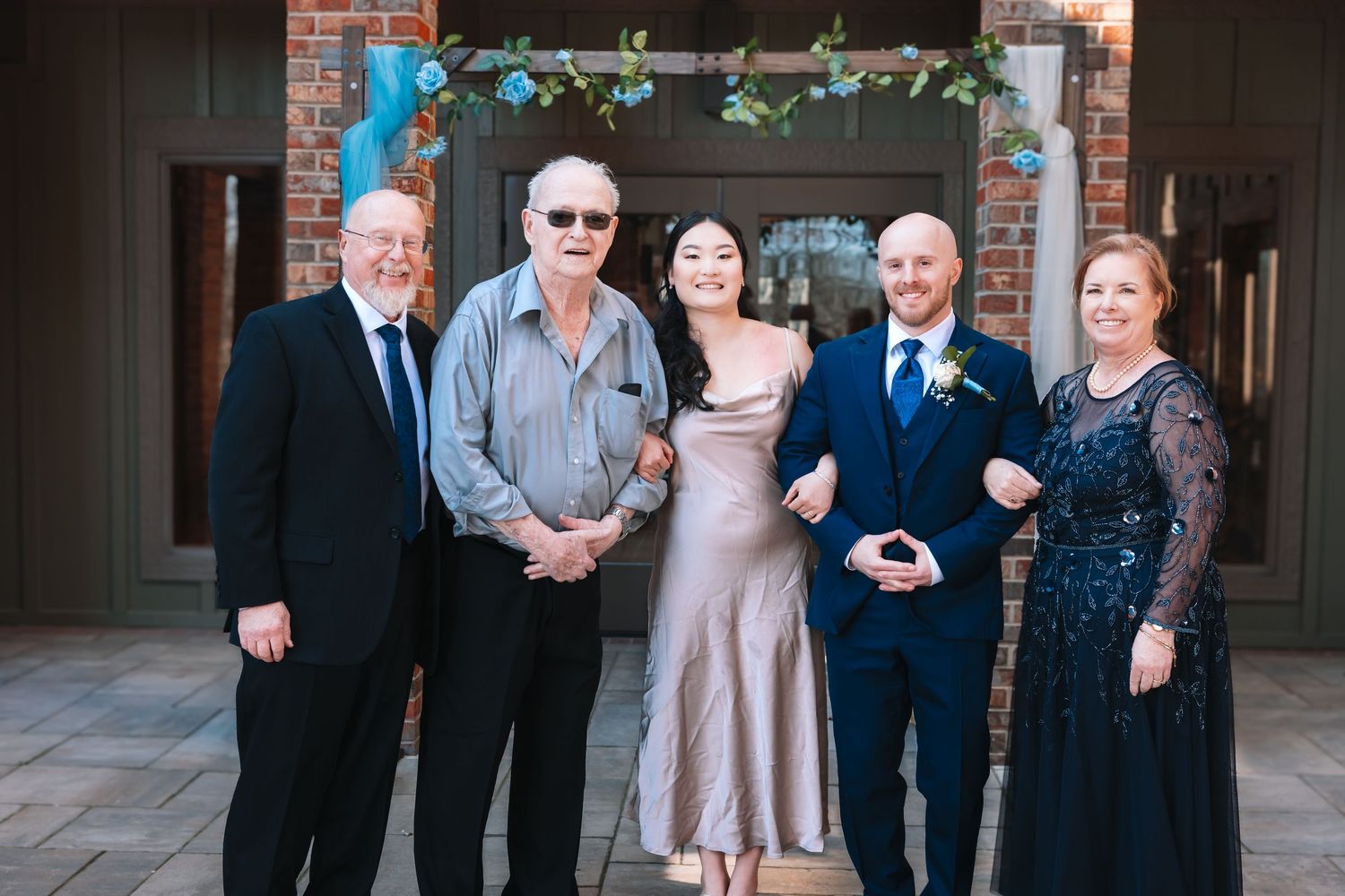 A wedding couple in elegant attire poses with three family members in front of a decorated doorway.