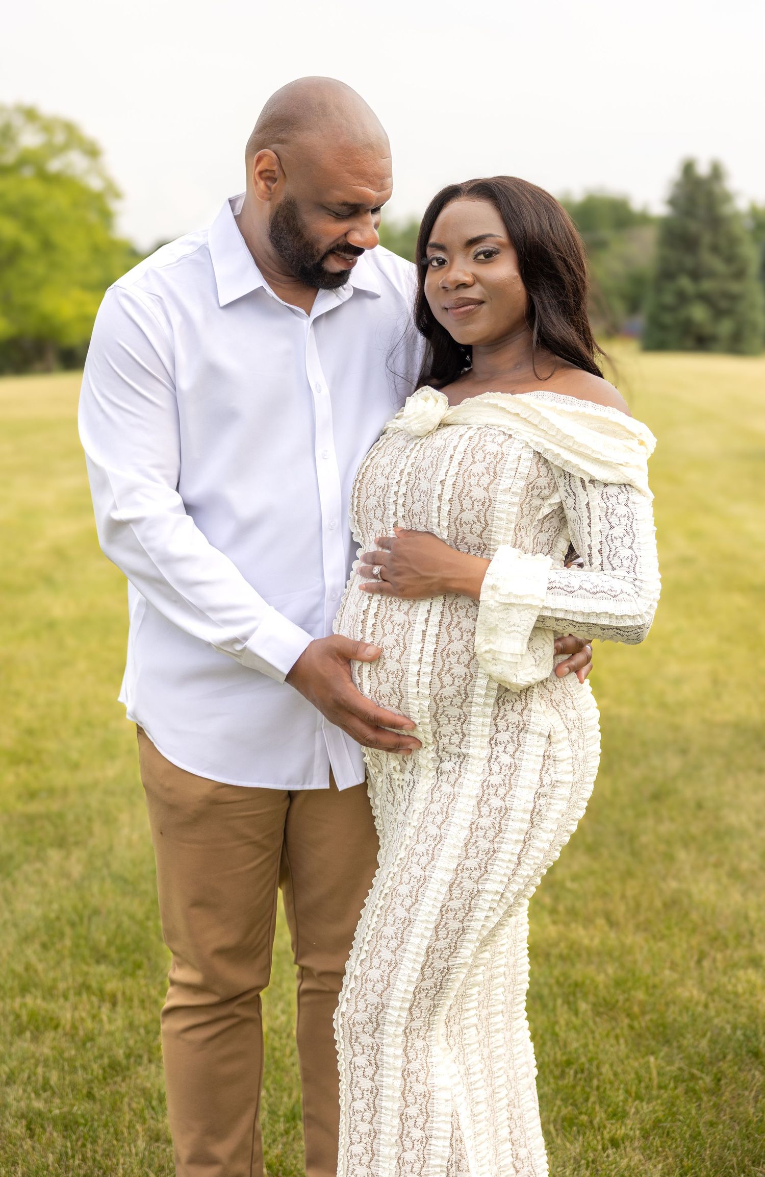 Expecting couple standing in green field, man embracing pregnant woman in white lace dress.
