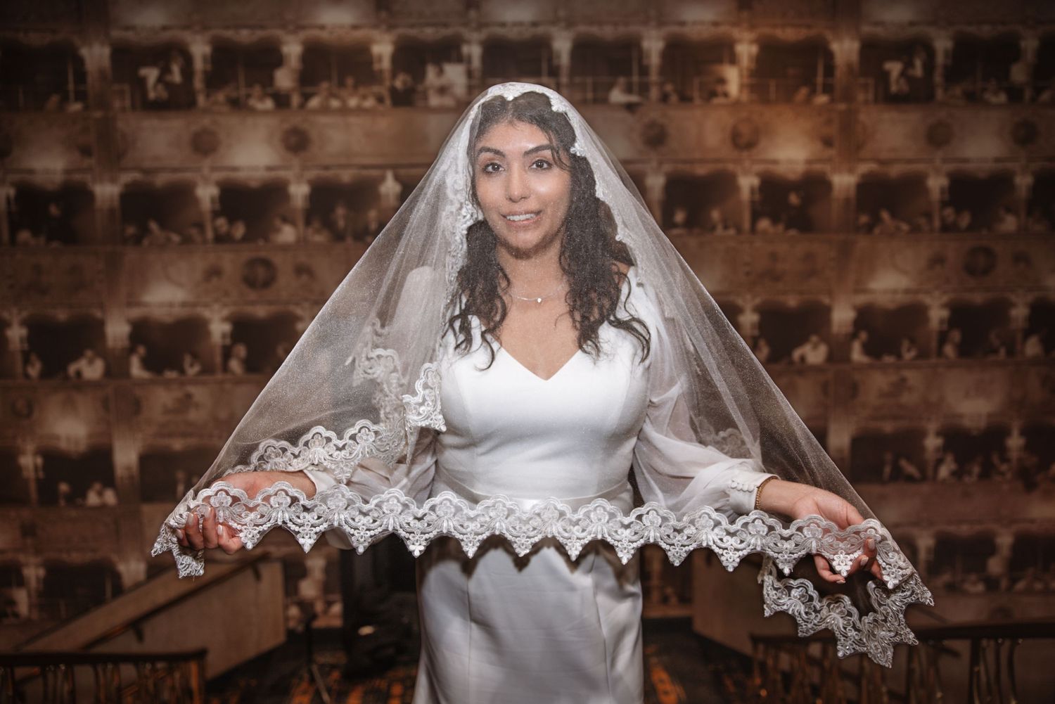 Bride in white satin dress spreads lace-edged veil wide in elegant venue.