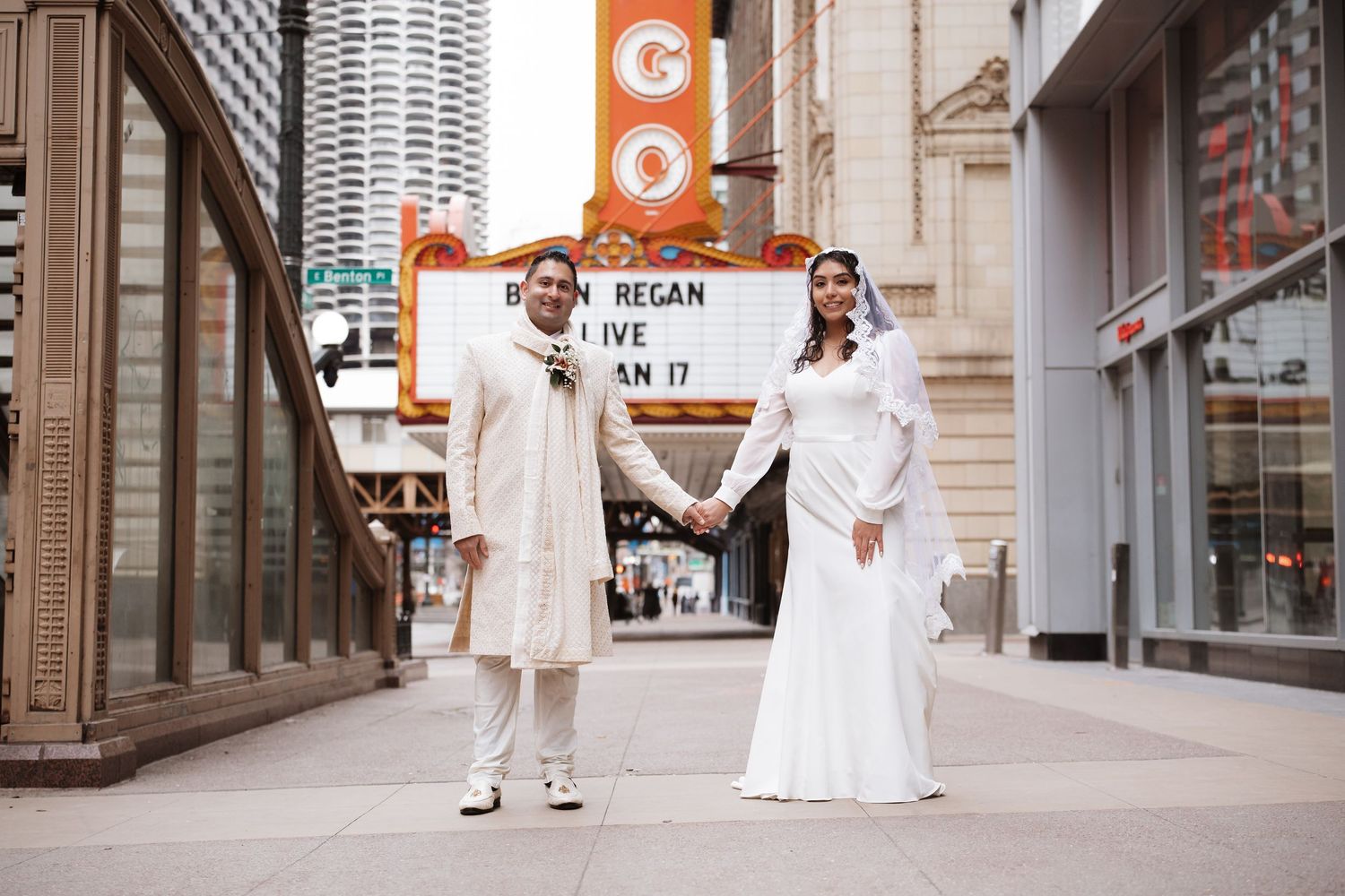 South Asian bride and groom in white wedding attire hold hands outside Chicago Theatre marquee.