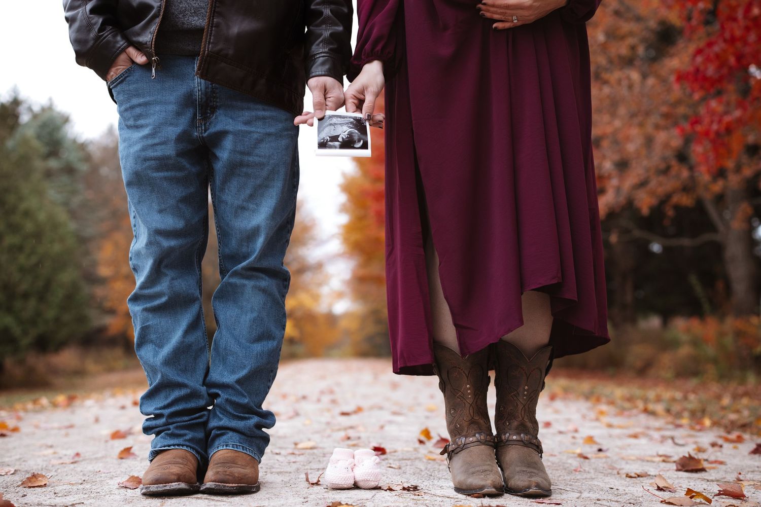 Couple on fall path holding ultrasound image with tiny baby shoes placed on ground between them.