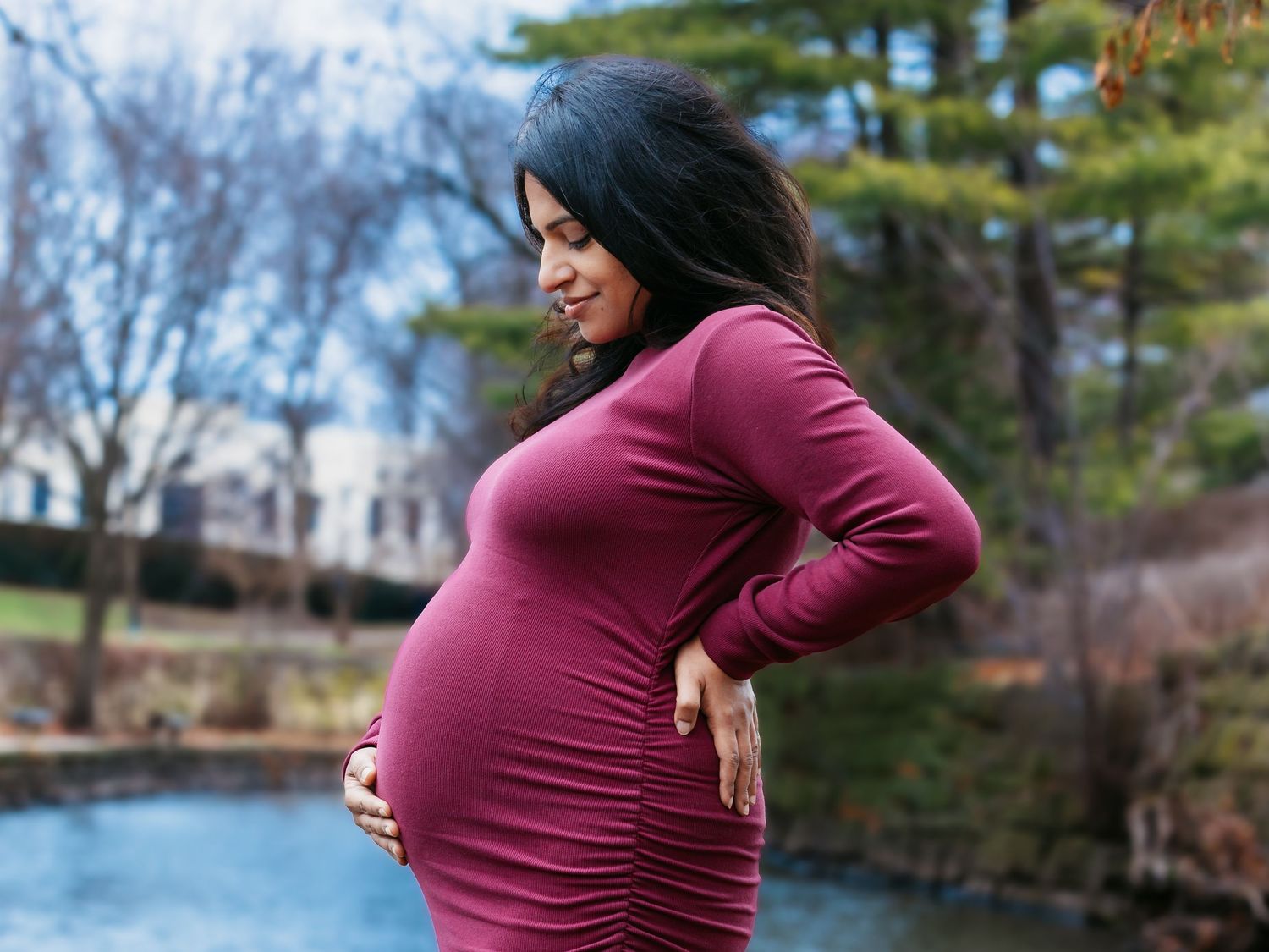 Pregnant woman in burgundy long-sleeve dress cradling baby bump outdoors by pond.