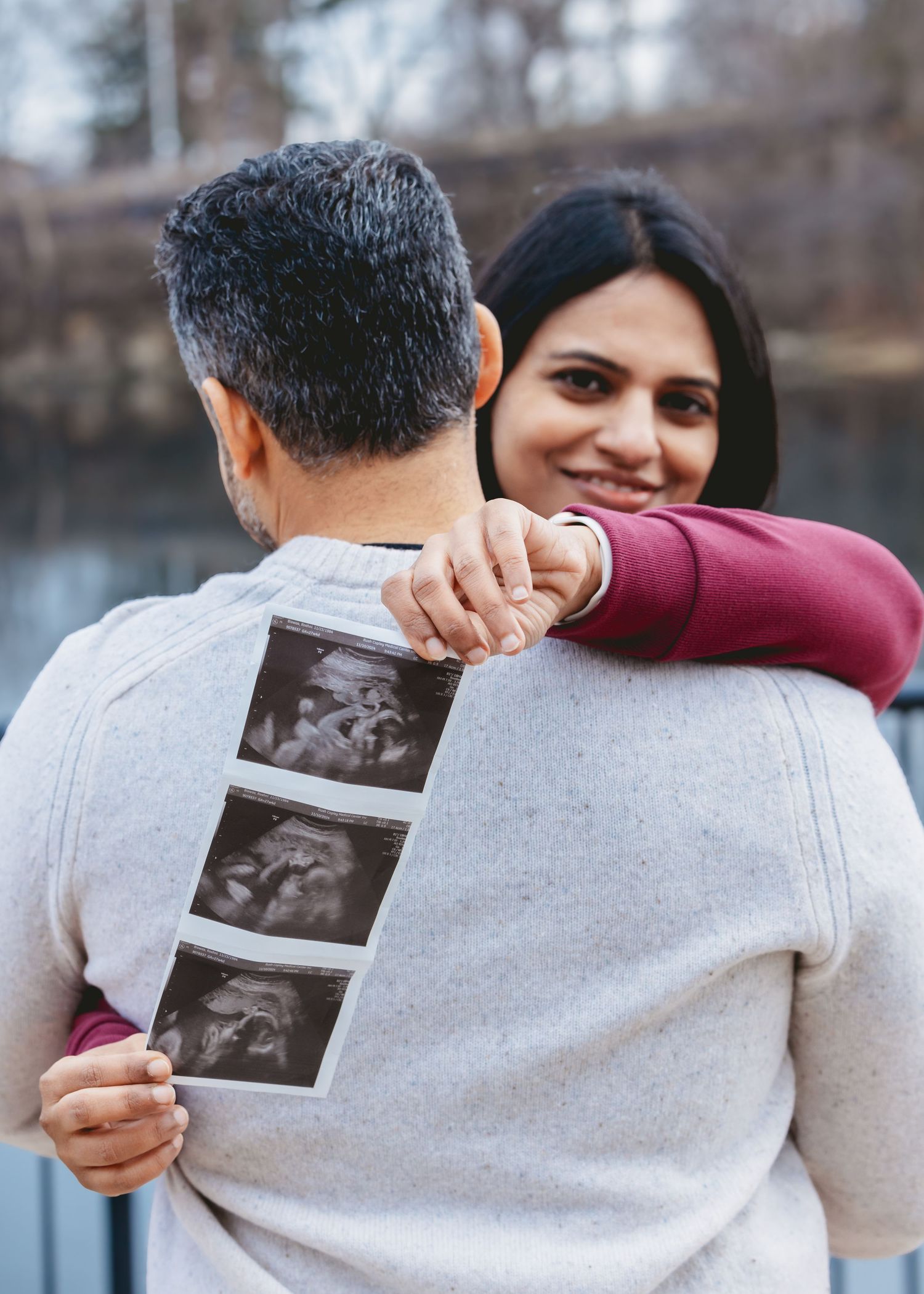 Couple sharing a tender embrace outdoors as man holds ultrasound photos, woman smiling happily.