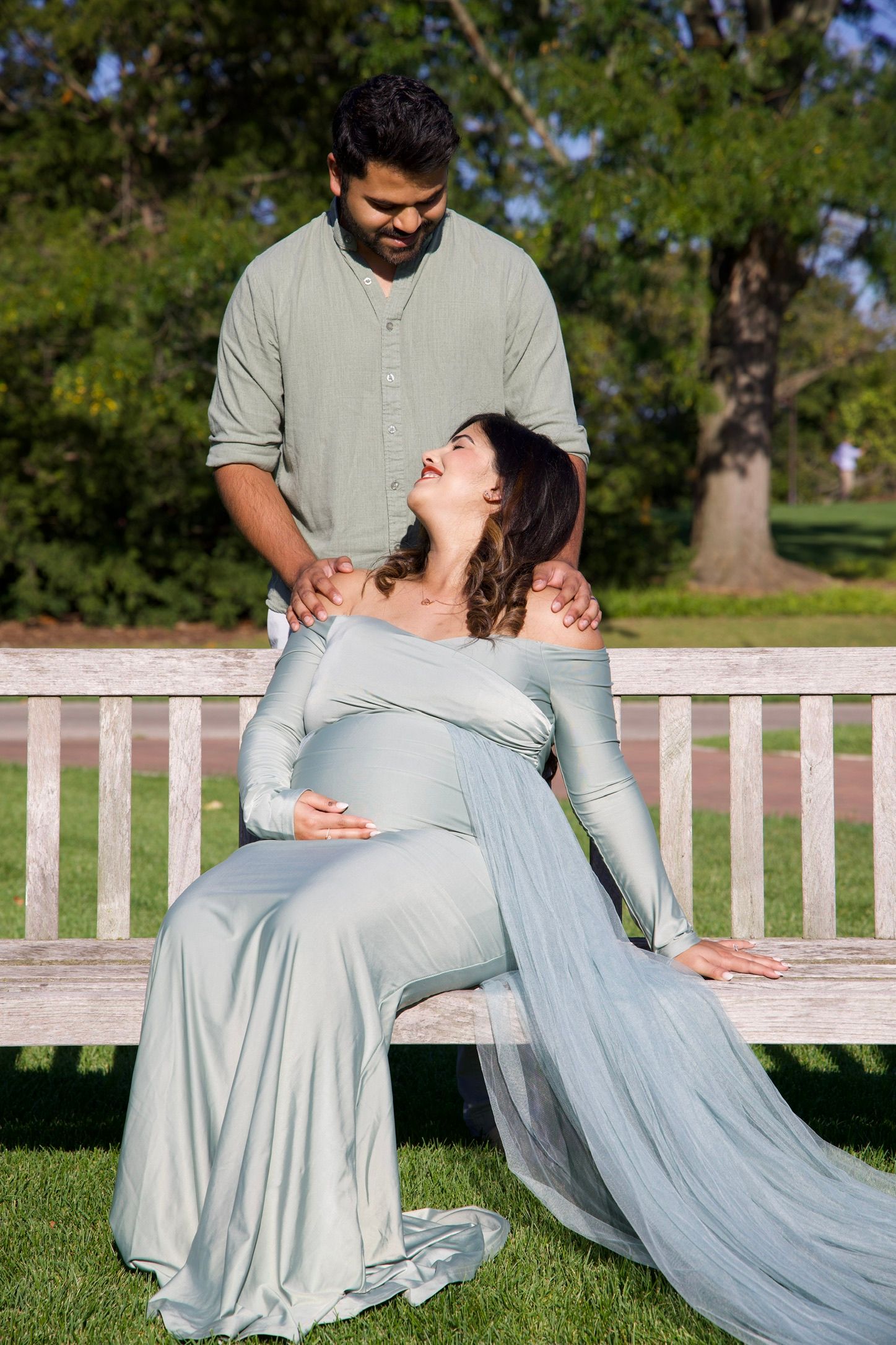Pregnant woman in flowing sage gown sitting on park bench laughing up at her partner standing behind her.