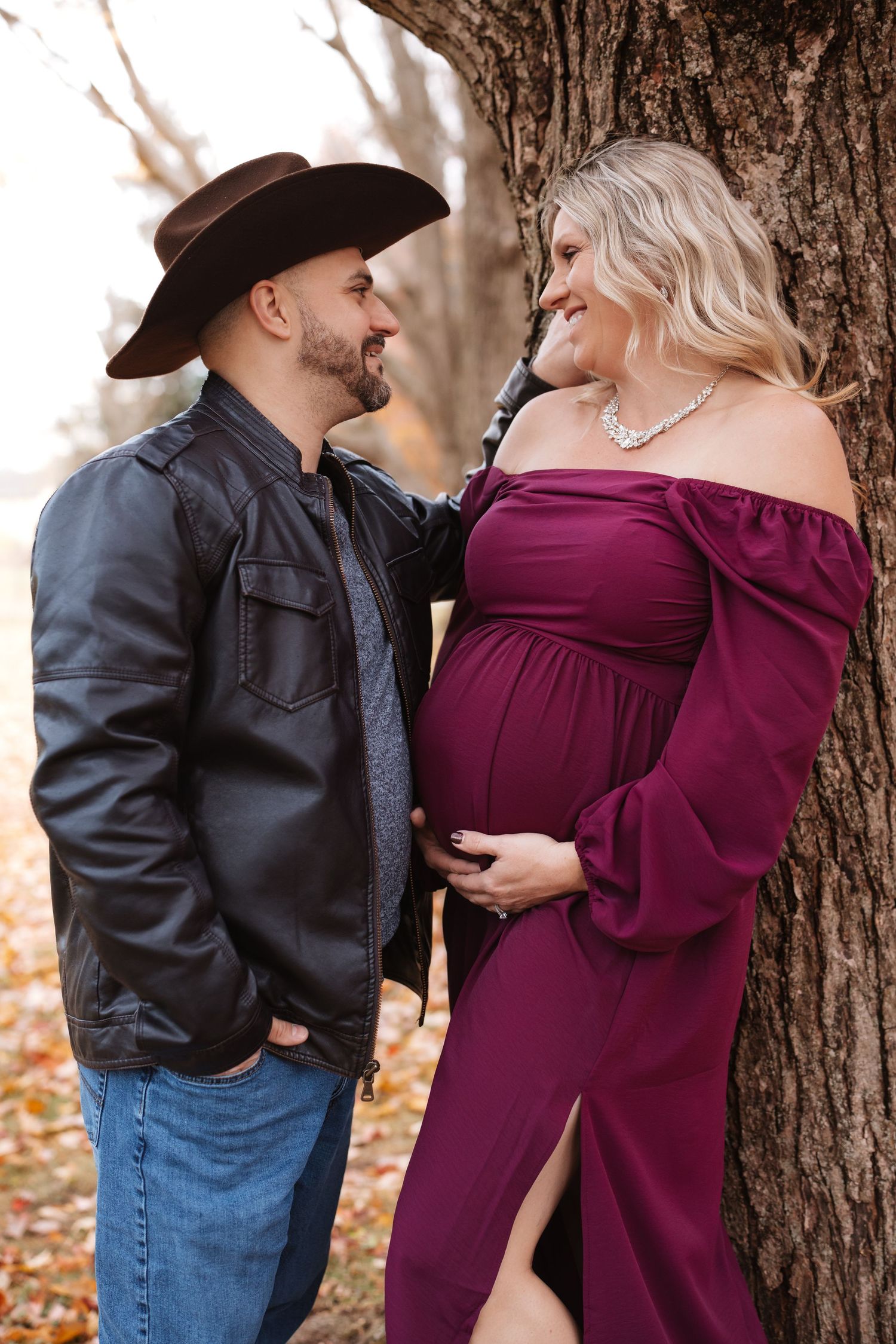 Couple gazing at each other in fall park, man in cowboy hat with pregnant woman in burgundy off-shoulder dress.