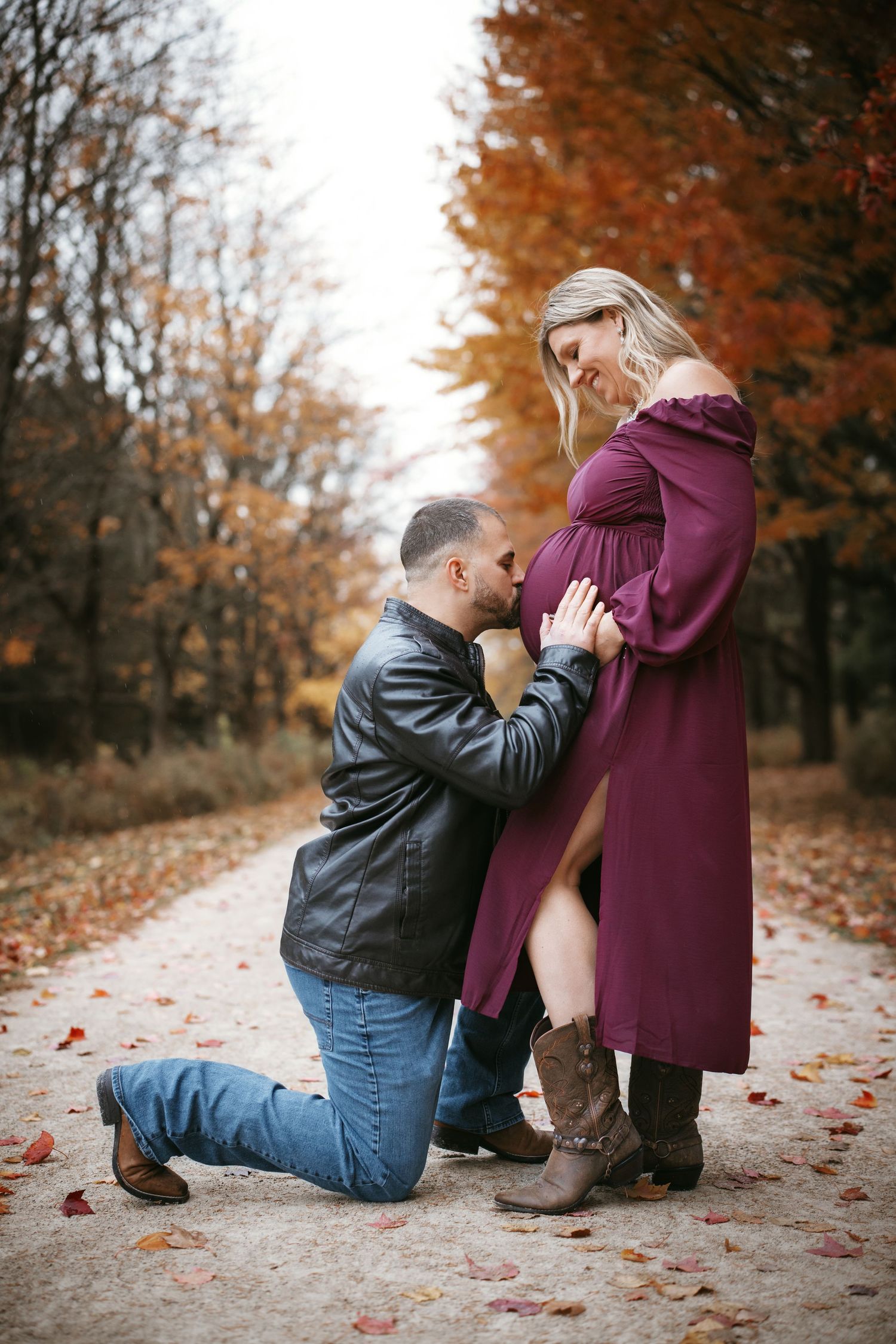 Couple sharing a tender moment on an autumn path, man kneeling to kiss pregnant woman's belly in purple dress.