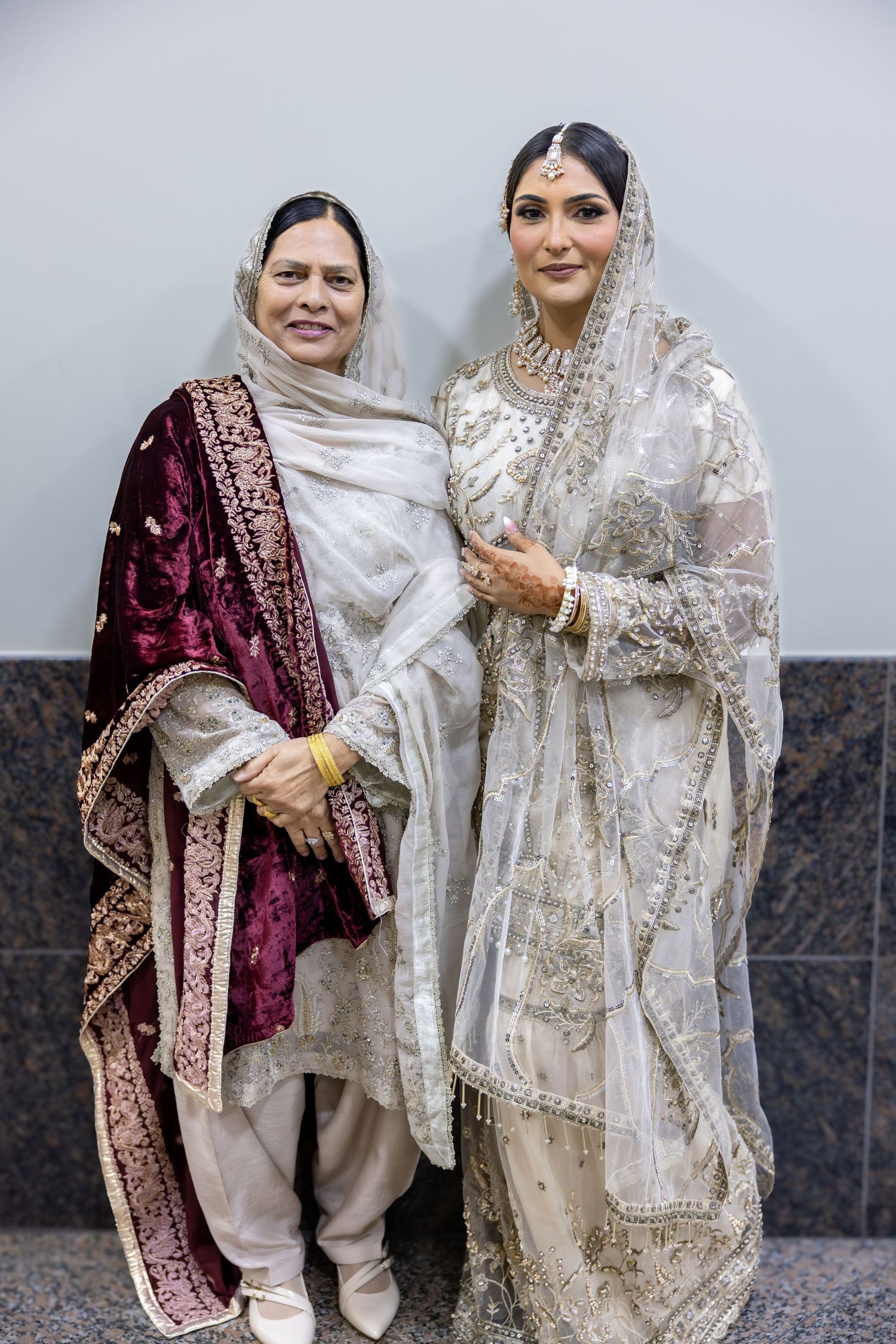 A South Asian bride in white embroidered bridal attire stands beside an older woman in a burgundy velvet salwar suit.