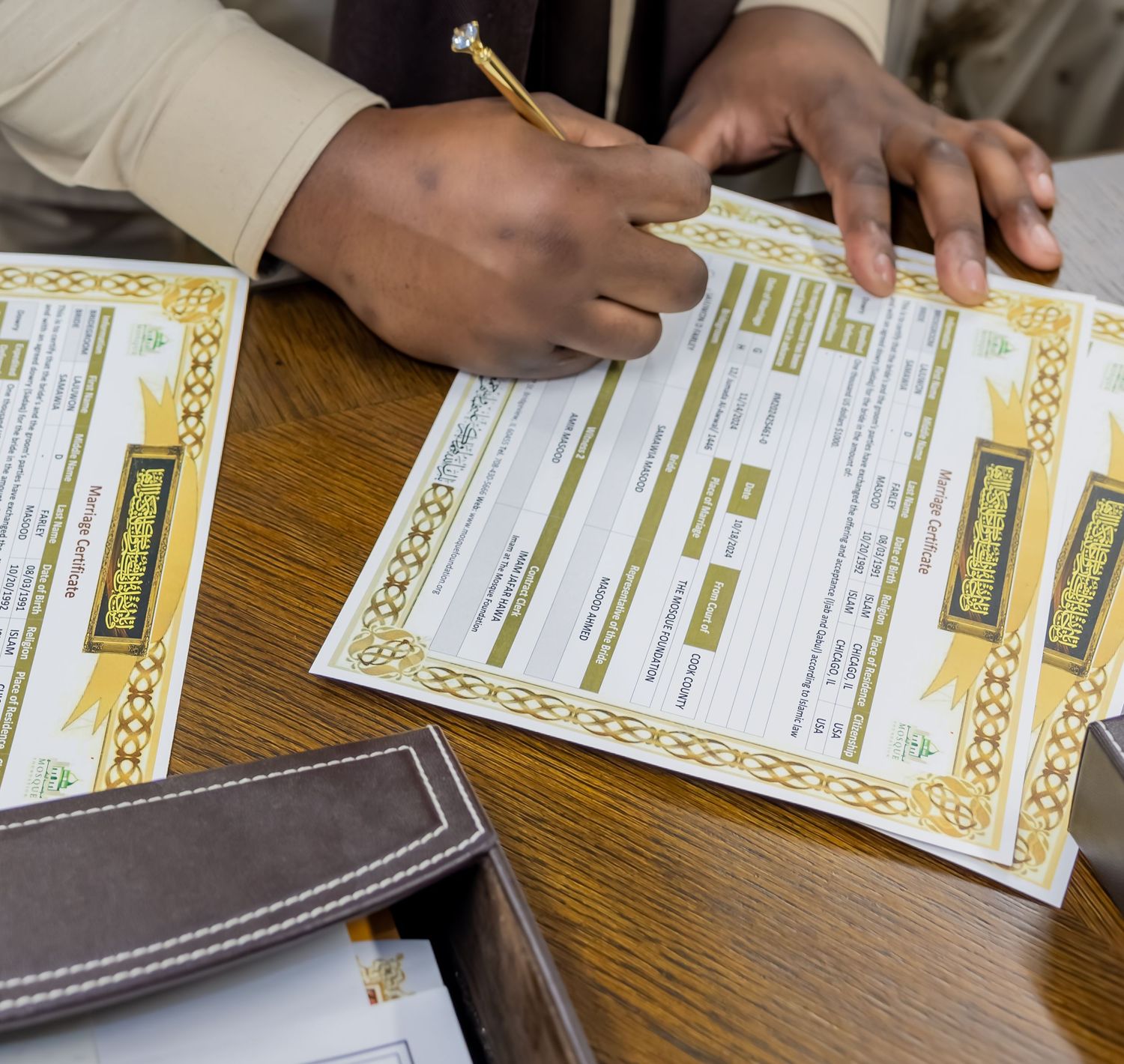 Hands signing ornate Islamic marriage certificate documents with gold decorative border on wooden desk.