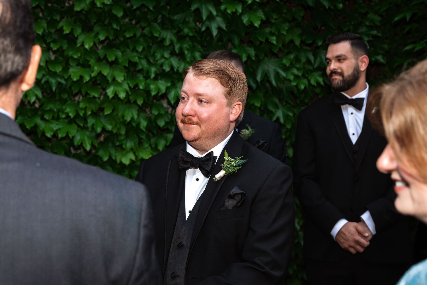 Groom wearing black bow tie tuxedo smiling joyfully at outdoor wedding ceremony with green ivy background.