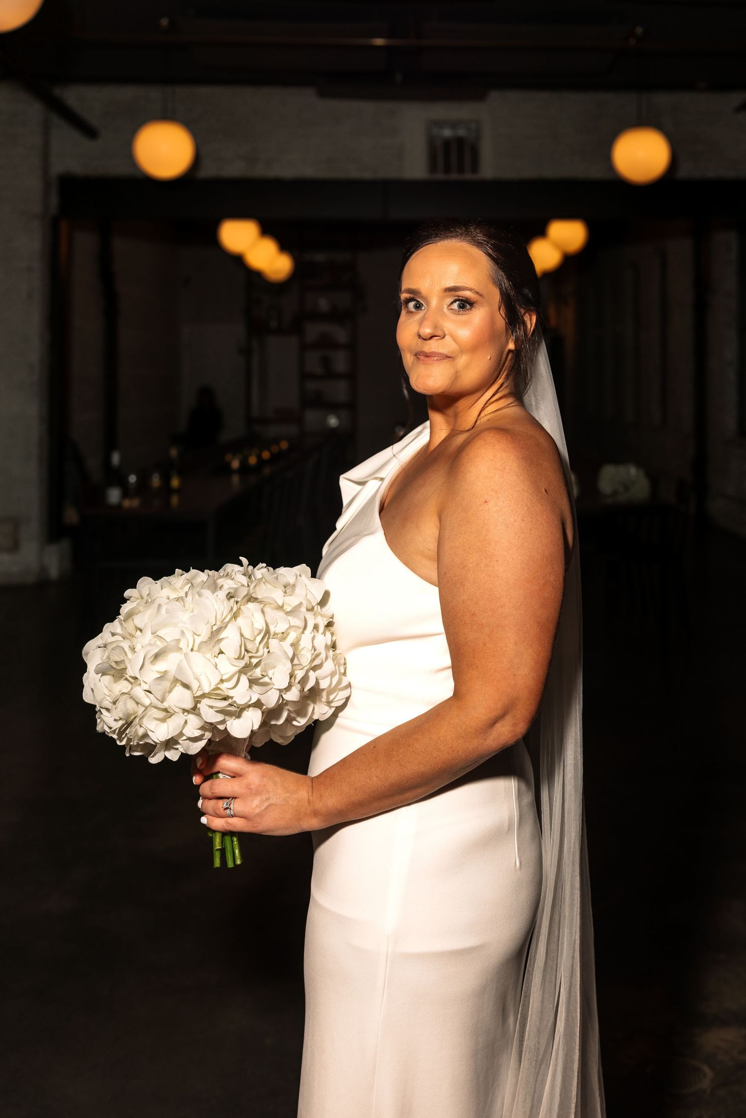 Elegant bride in sleek white gown holding white hydrangea bouquet with veil in dimly lit venue.
