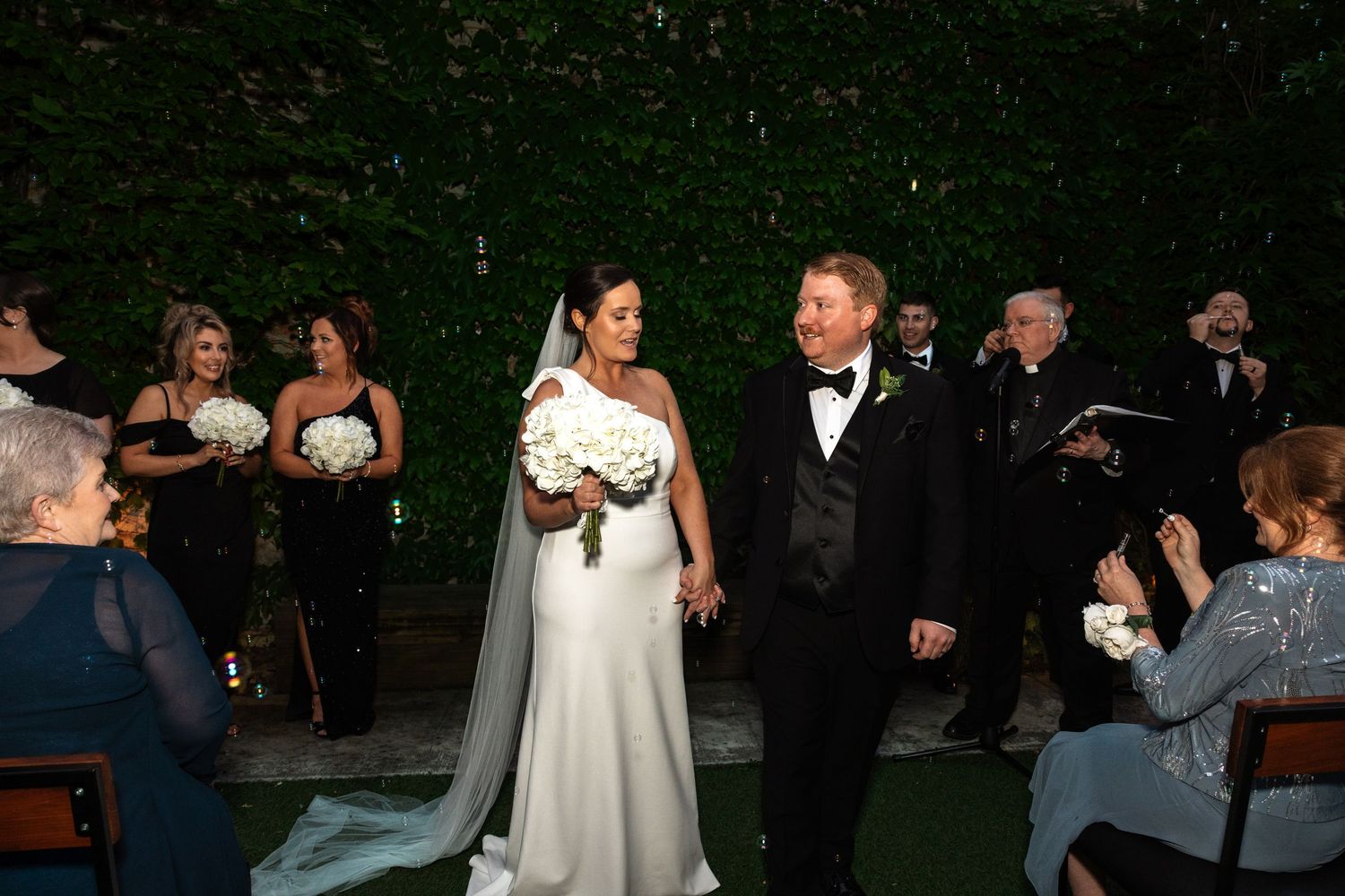 Bride holds bouquet as newlyweds walk back down aisle at outdoor evening wedding.