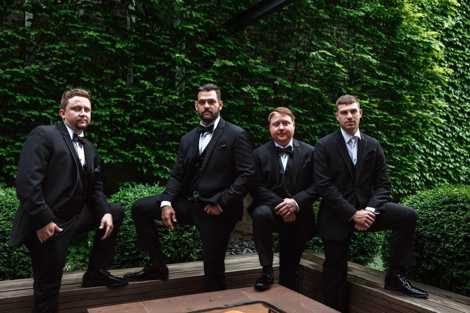 Four groomsmen in black tuxedos seated and posing in front of a green ivy wall.