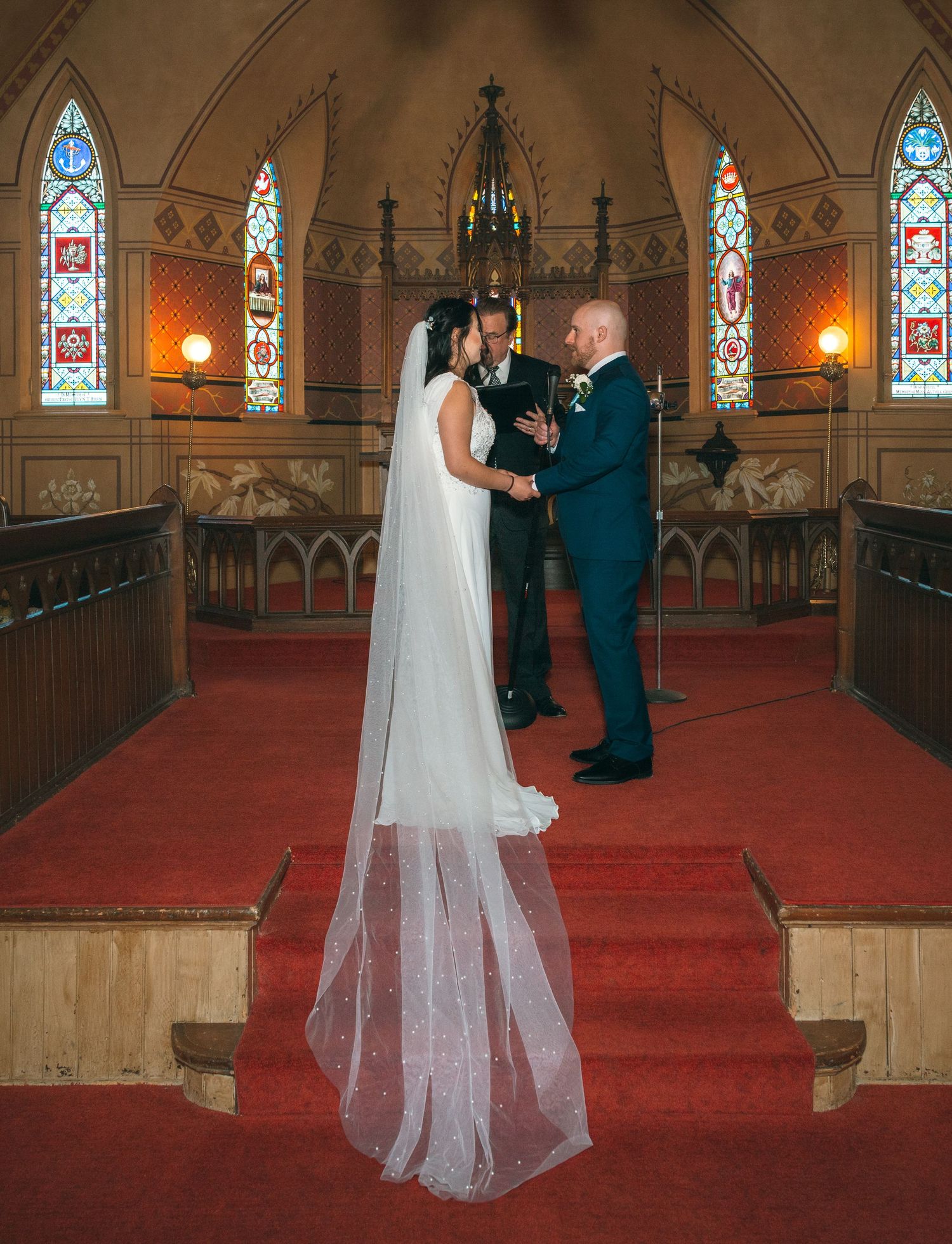Bride in white gown with long veil and groom in navy suit exchange vows at church altar.